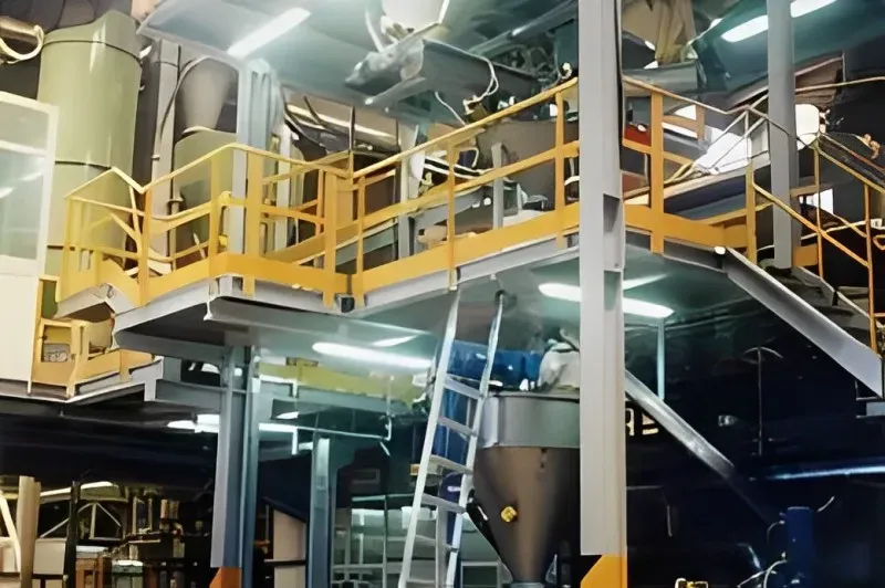 Piattaforma industriale con ringhiera gialla, macchinari grigi e argento. Una scala conduce alla salita.