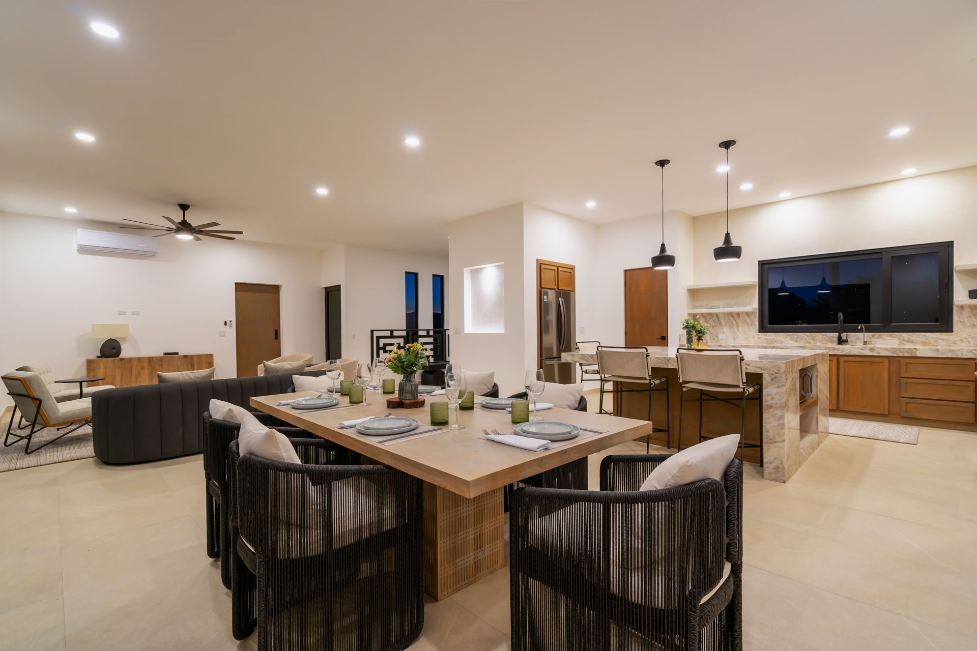 A modern, open-concept living and dining room with a large wooden table, rope-style chairs, and a kitchen island.
