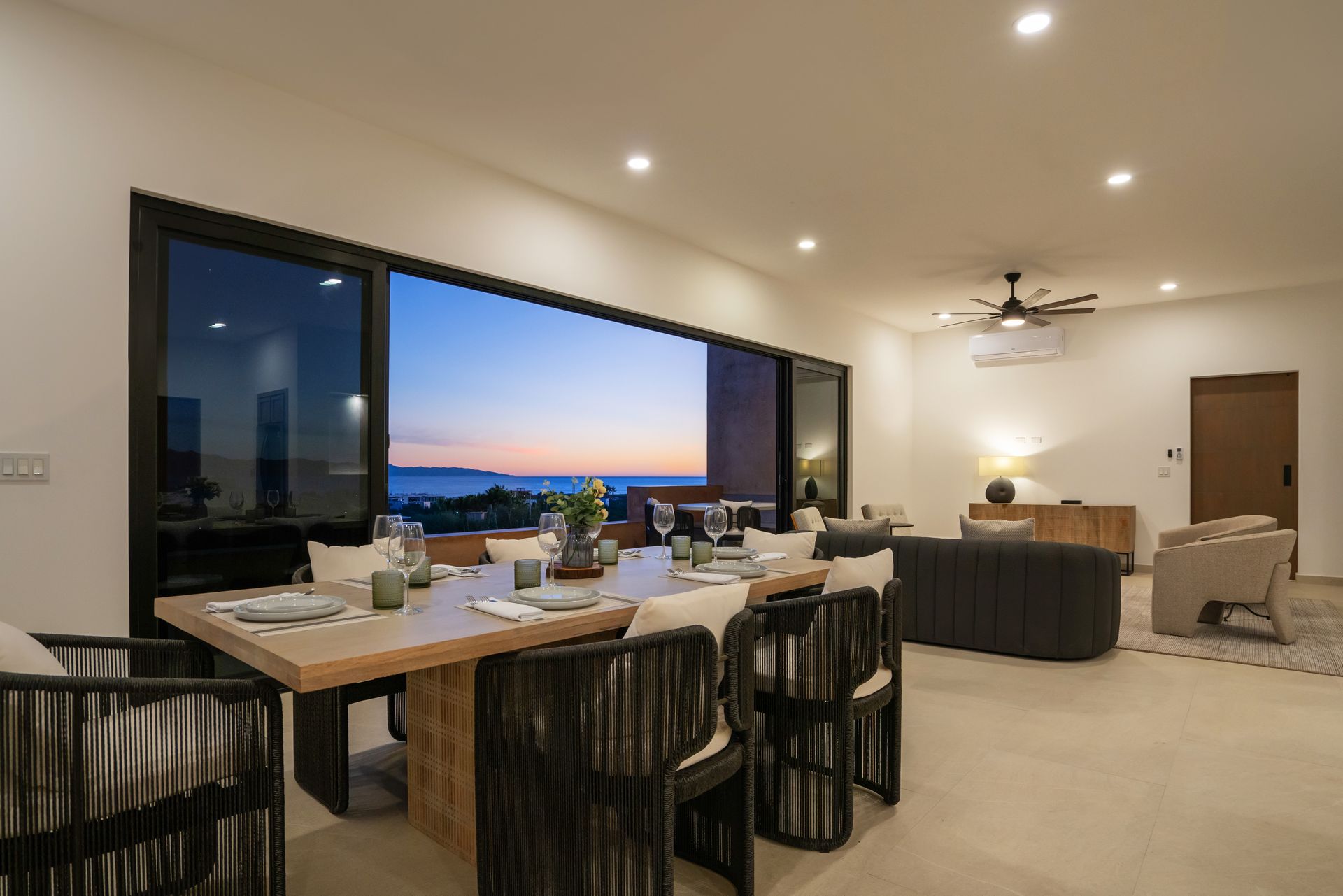 An indoor dining area flows into an open-plan living room with large glass doors showing a sunset view over the ocean.