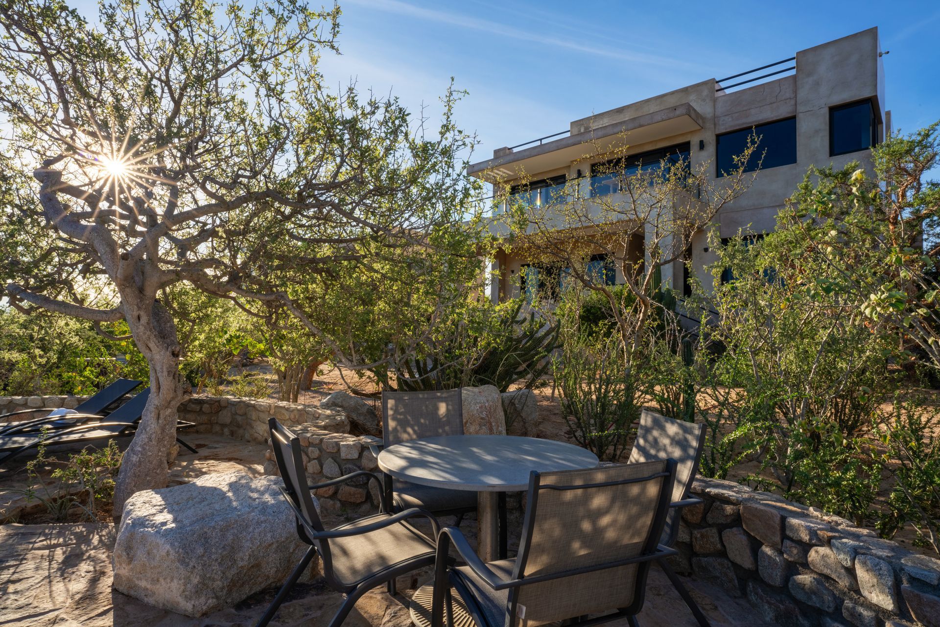 A patio with a round table and chairs sits in front of a modern desert-style home under a bright, sunny sky.