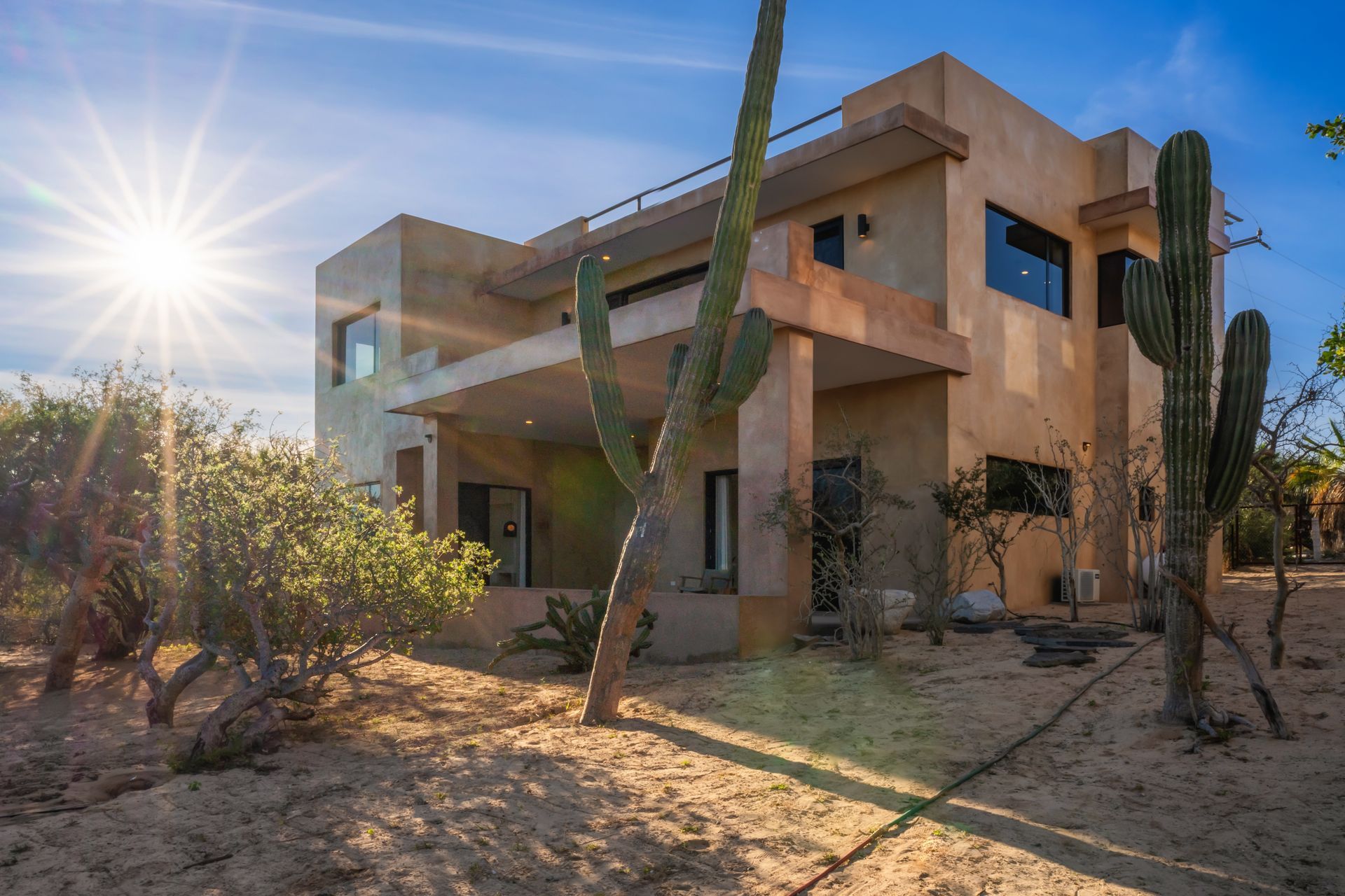 A multi-story beige stucco house under a bright sun, surrounded by desert landscape and tall cacti.