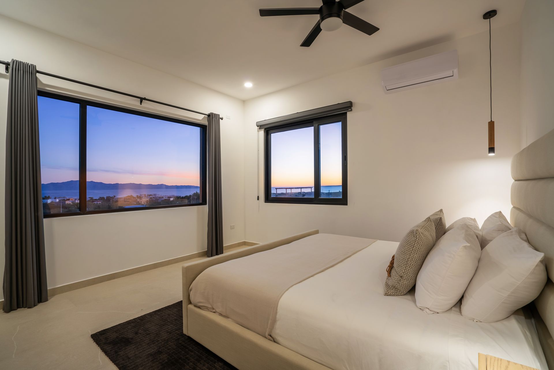A modern, bright bedroom with a queen bed, grey curtains, and two large windows overlooking a scenic coastal sunset.