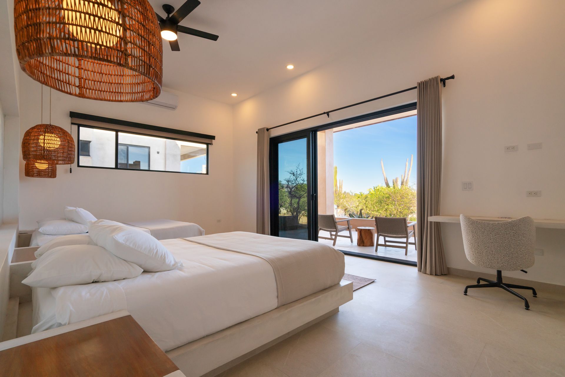 A modern, minimalist bedroom with two beds, woven pendant lights, and a large glass door leading to a sunny patio.