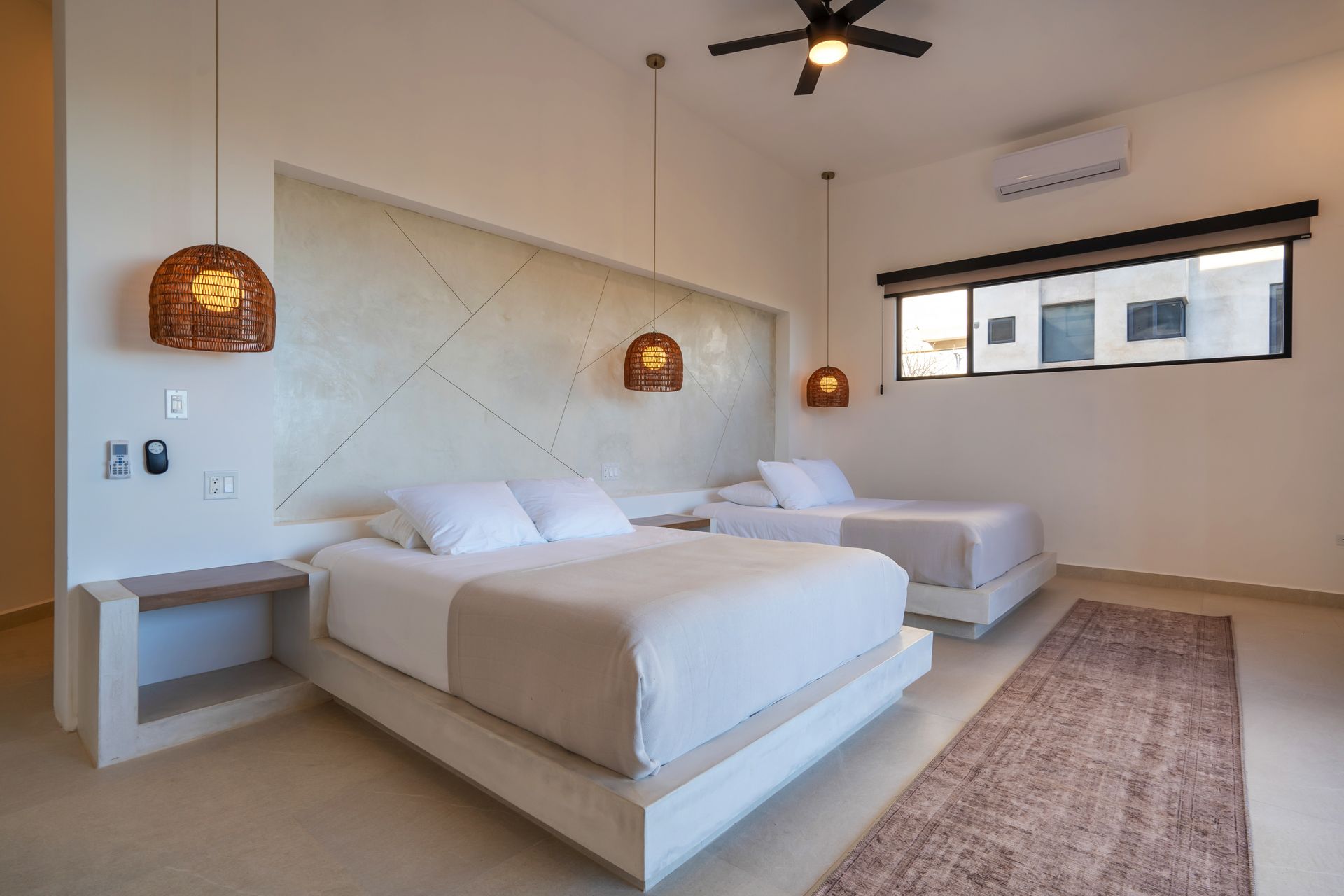 A modern bedroom with two queen beds, minimalist decor, beige walls, and warm hanging lights.