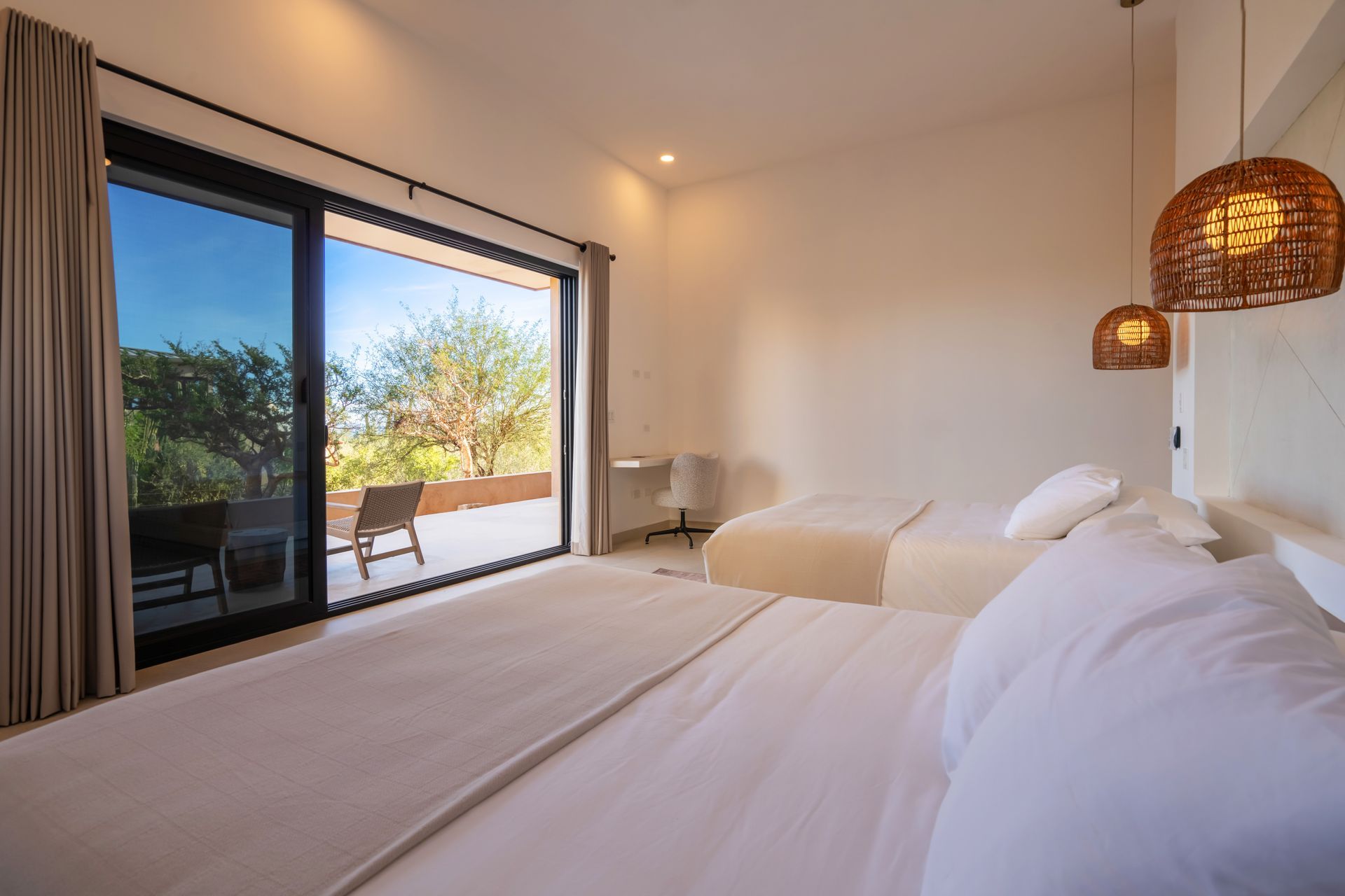 A minimalist bedroom with two beds, neutral bedding, woven pendant lights, and a sliding glass door opening to a terrace.