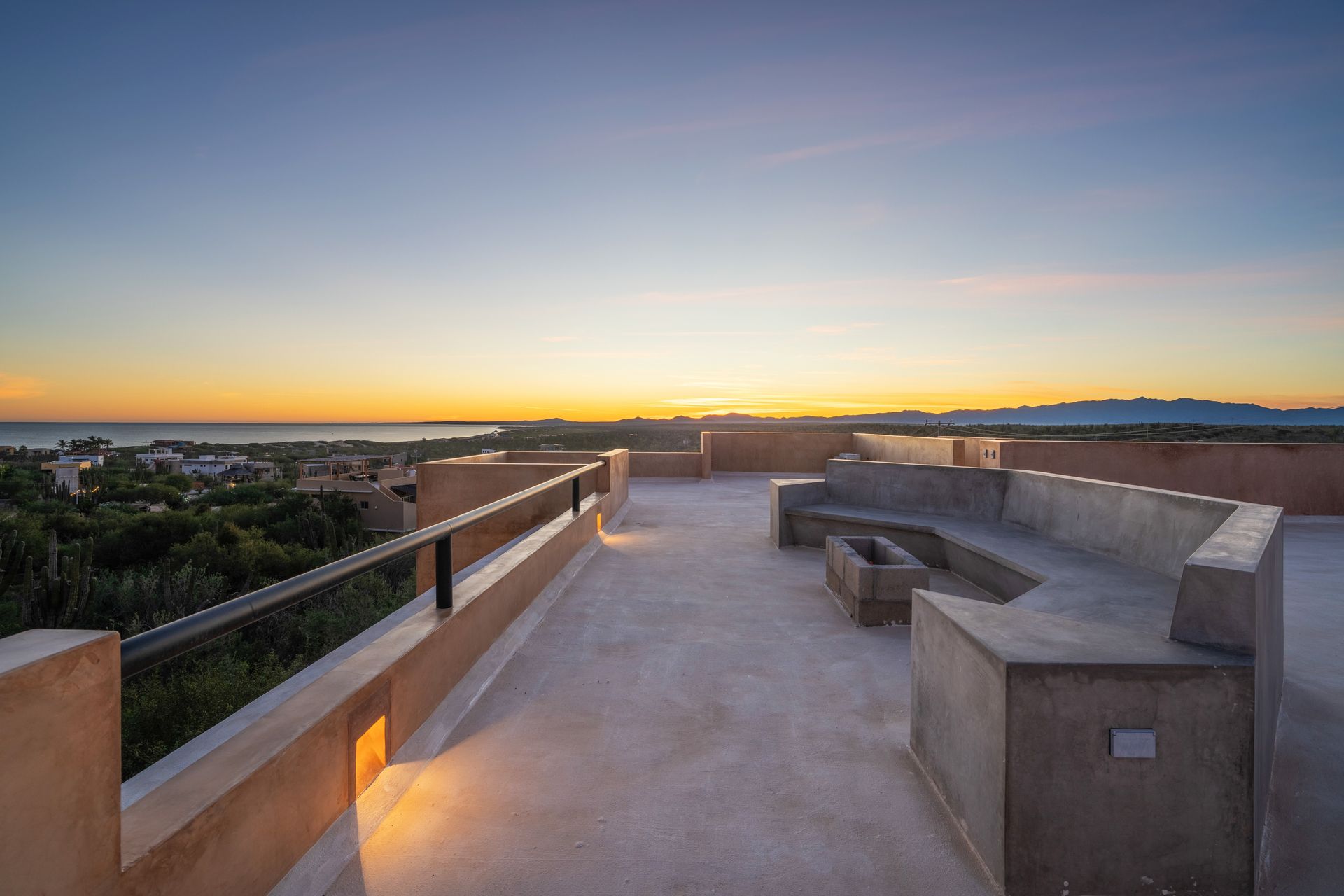 A rooftop terrace with concrete benches and a fire pit at sunset overlooking a landscape and distant ocean.