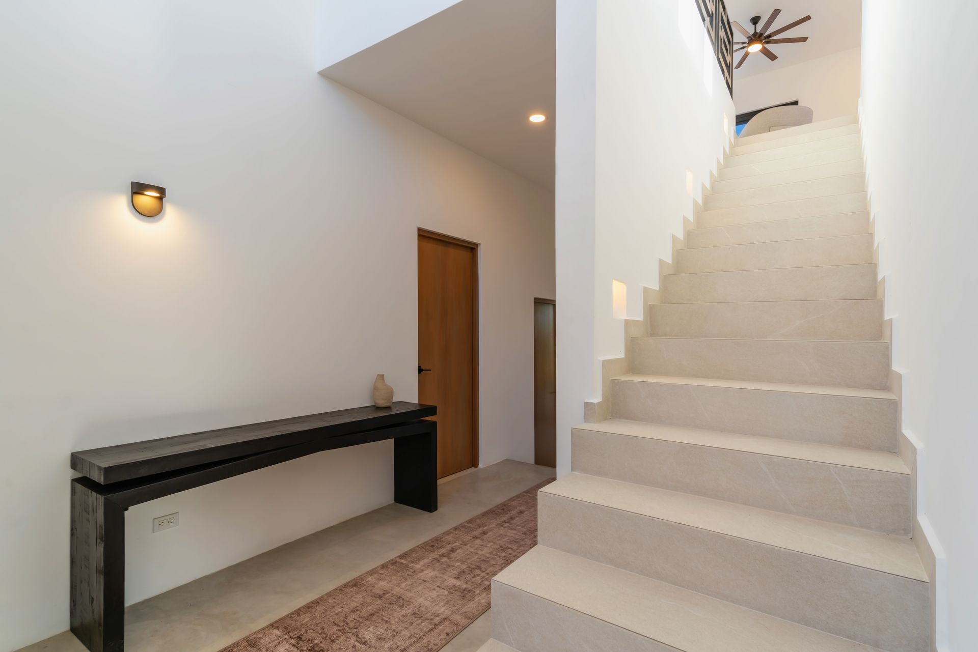 A minimalist hallway with a long black console table, a wooden door, and a wide light-toned staircase leading upward.