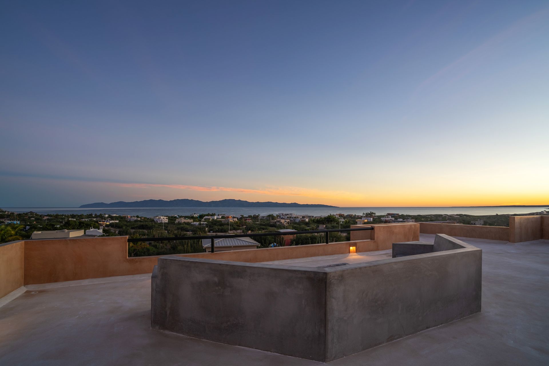 A concrete rooftop terrace overlooks a coastal town and distant island during a calm, golden-hued sunset.