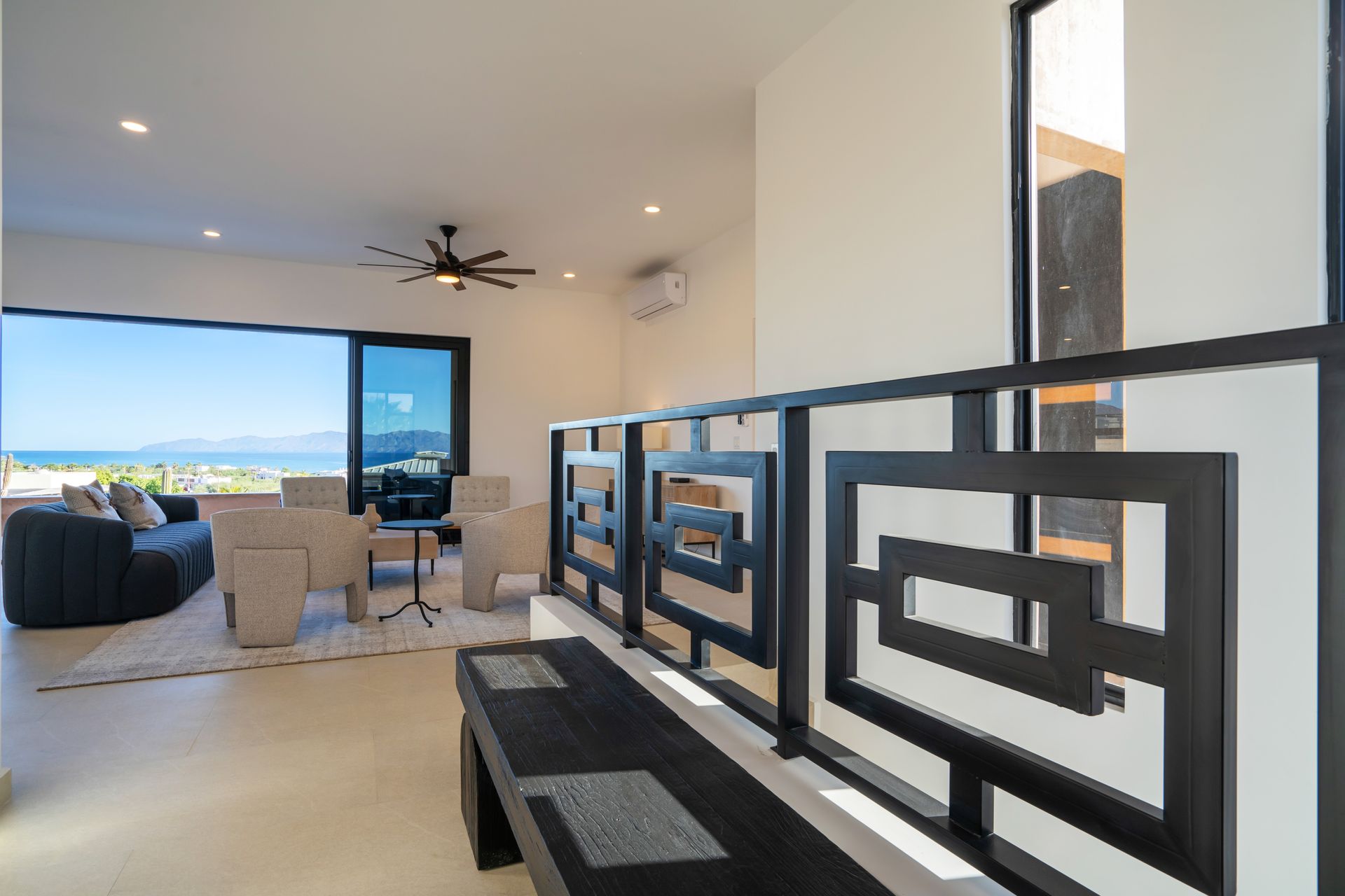 Modern interior with a black geometric staircase railing overlooking a living area and balcony with an ocean view.