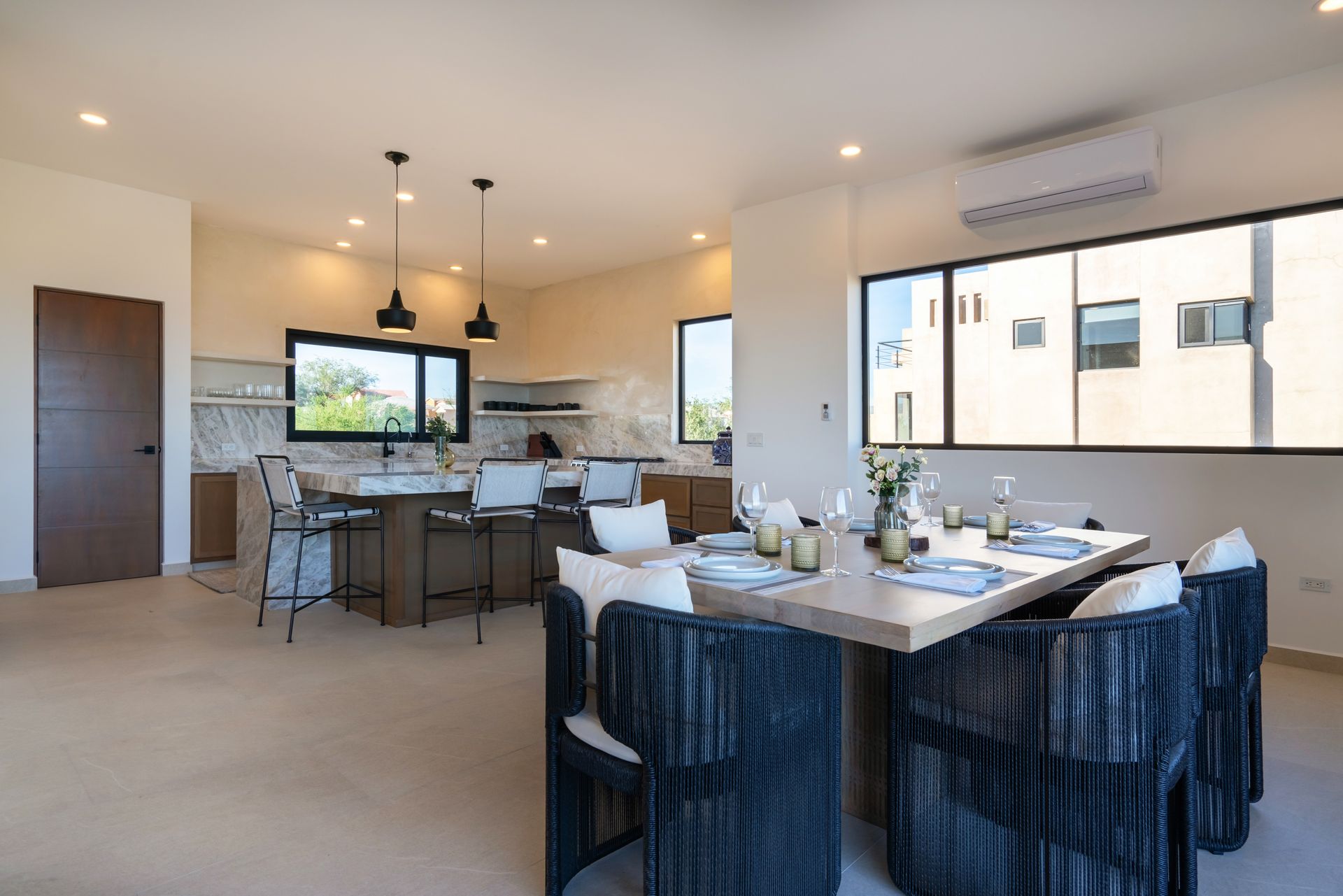 A modern, open-concept dining area and kitchen with a wooden dining table, dark wicker chairs, and a kitchen island.