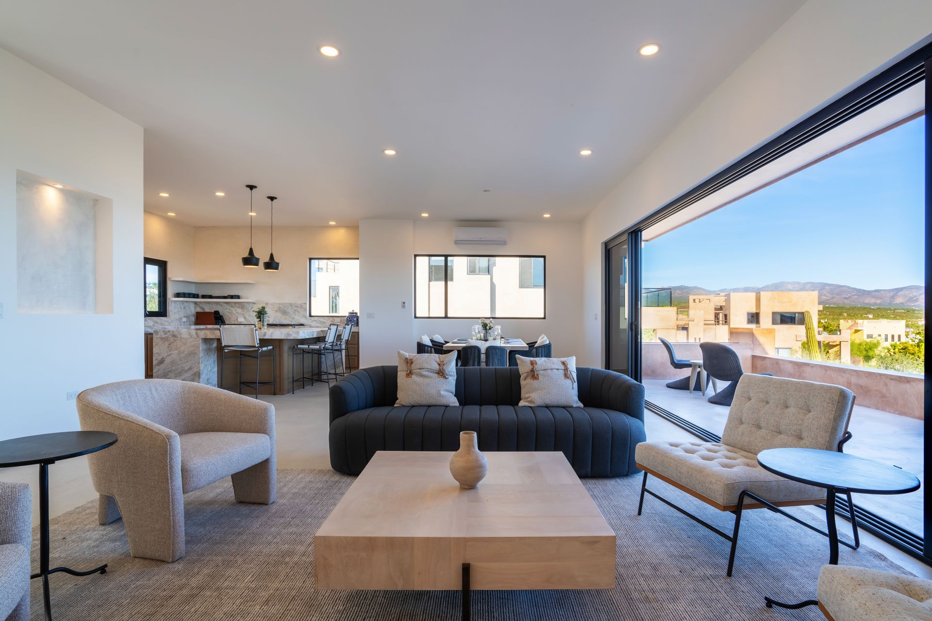 Modern living room with a gray sofa, two cream accent chairs, light wood coffee table, and large sliding glass wall.