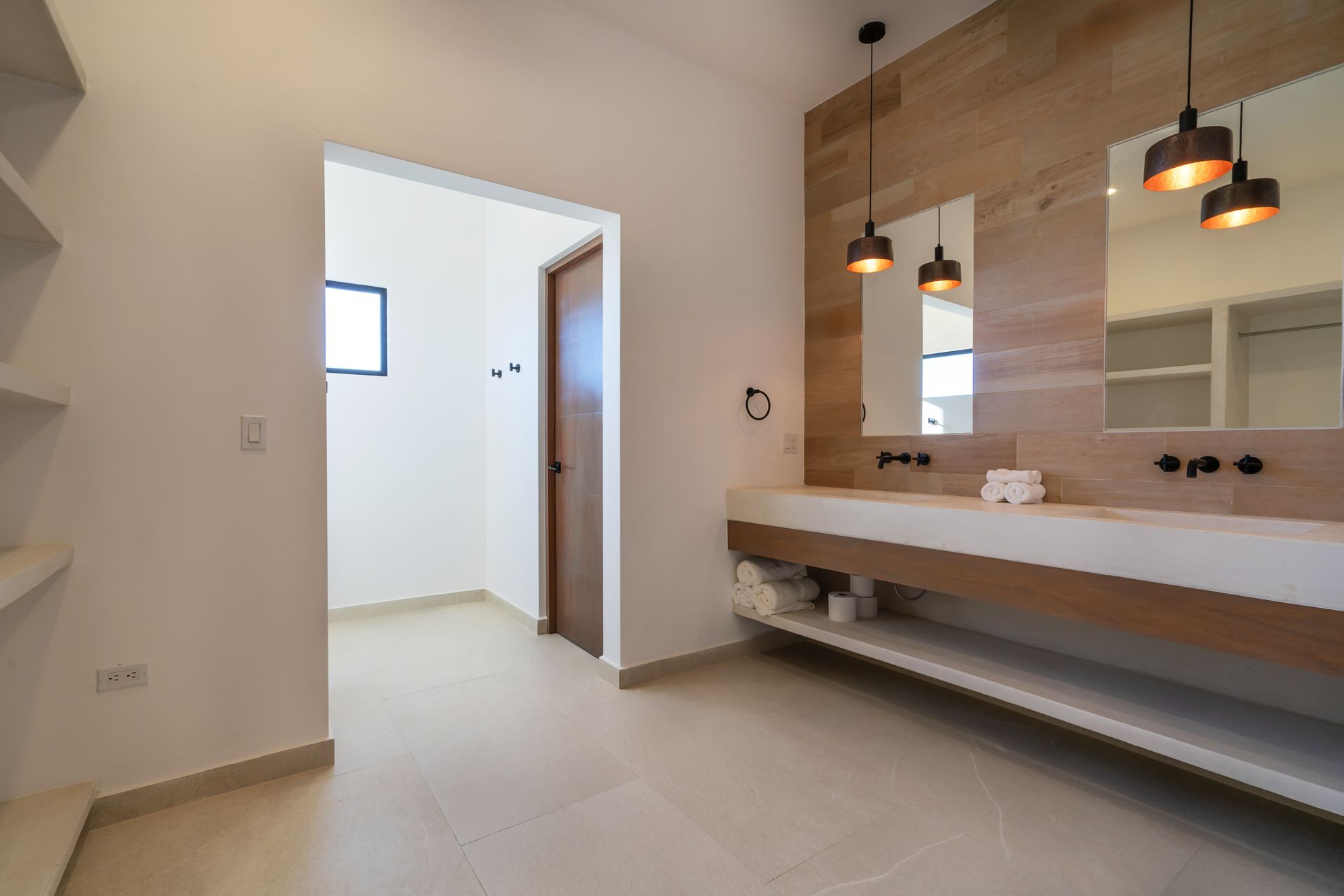 Modern bathroom with a double sink vanity, wood-paneled wall, hanging pendant lights, and an open doorway to another room.