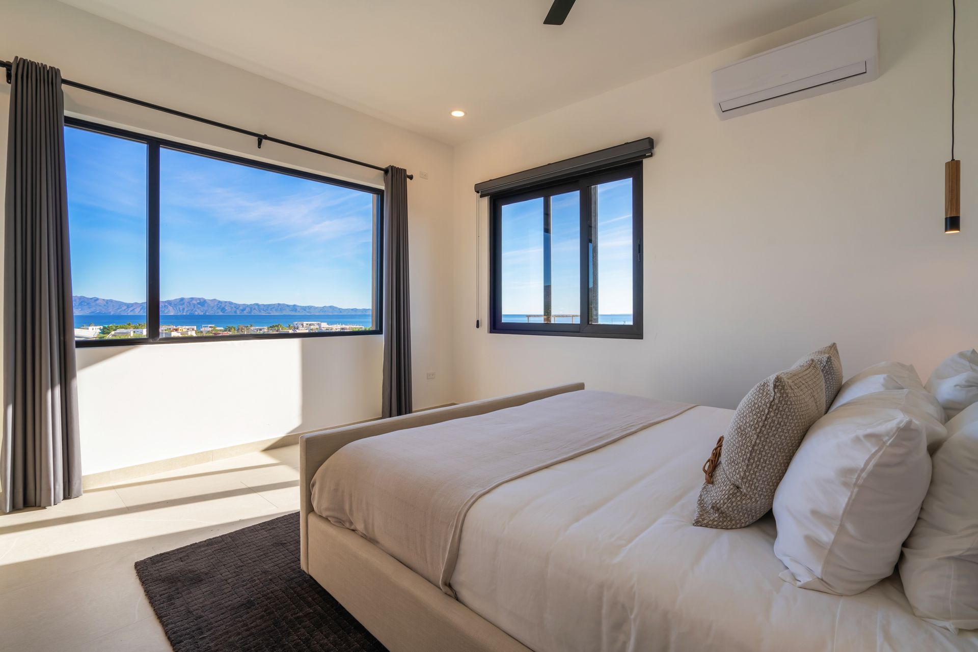A modern, minimalist bedroom with a large bed, neutral bedding, and two windows offering an expansive ocean view.