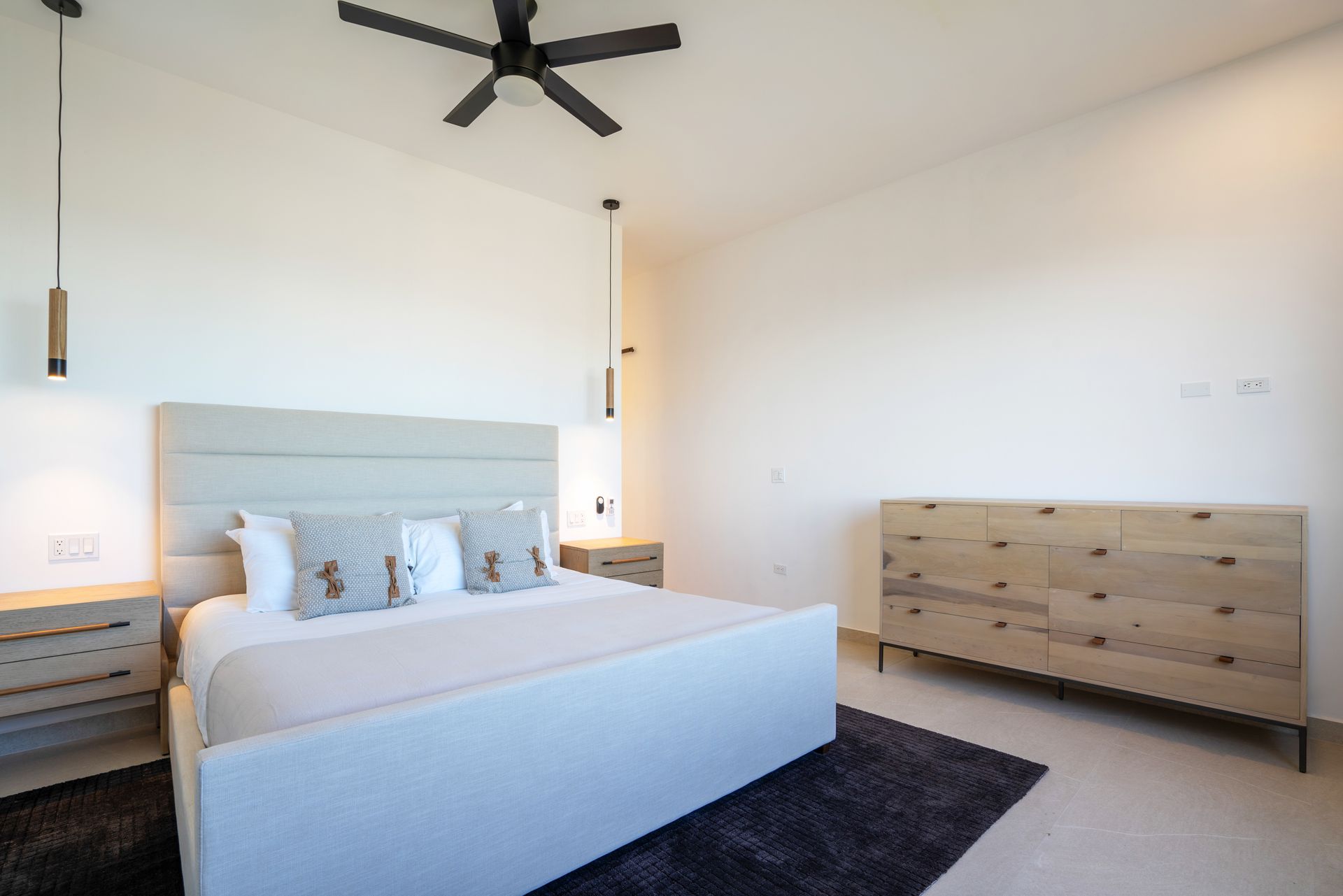 A modern, minimalist bedroom with a grey upholstered bed, matching bedside tables, a wooden dresser, and a ceiling fan.