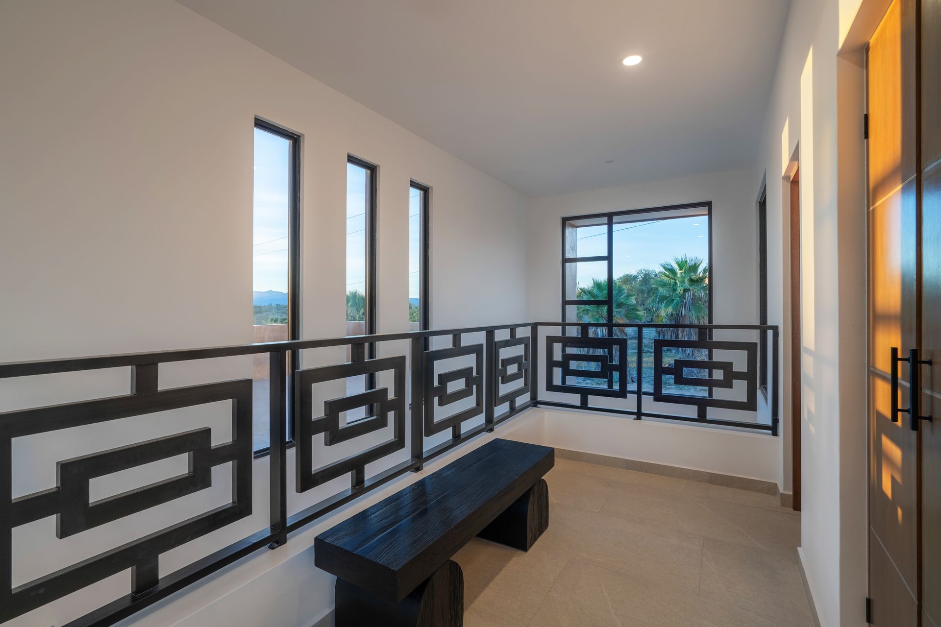 Modern landing area with white walls, a black wooden bench, a decorative metal railing, and windows overlooking greenery.