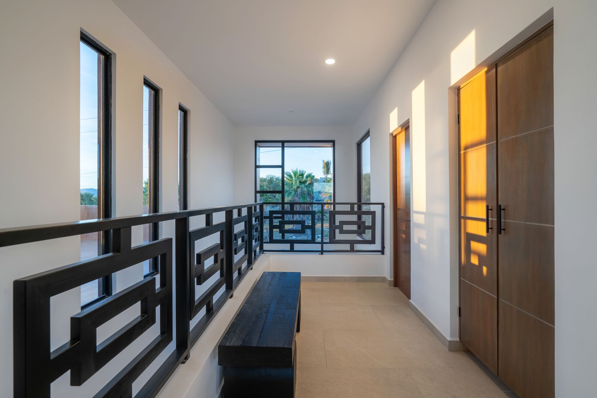 A sunlit indoor landing with white walls, a geometric black railing, a dark bench, and wooden doors.