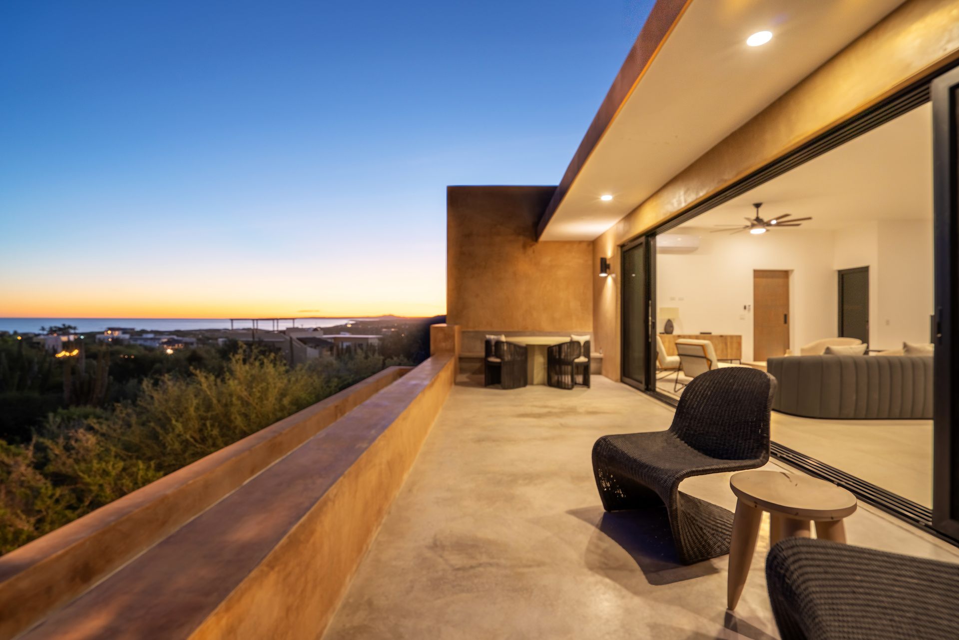 A balcony with wicker furniture overlooks a coastal town and the sea at sunset, adjacent to a living room with glass doors.