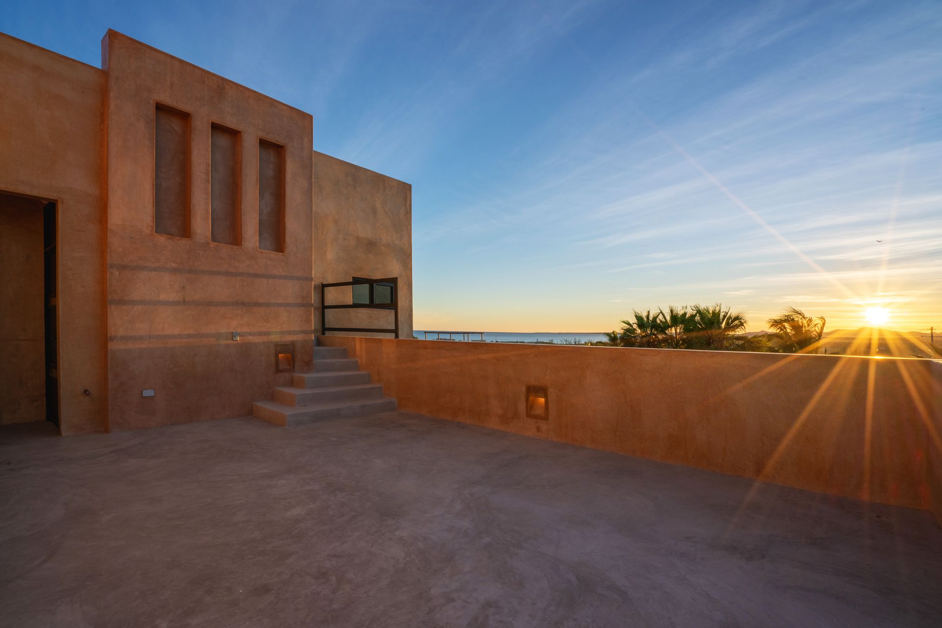 A spacious tan rooftop terrace at sunset with a low stone wall, stairs, and a clear view of the horizon.