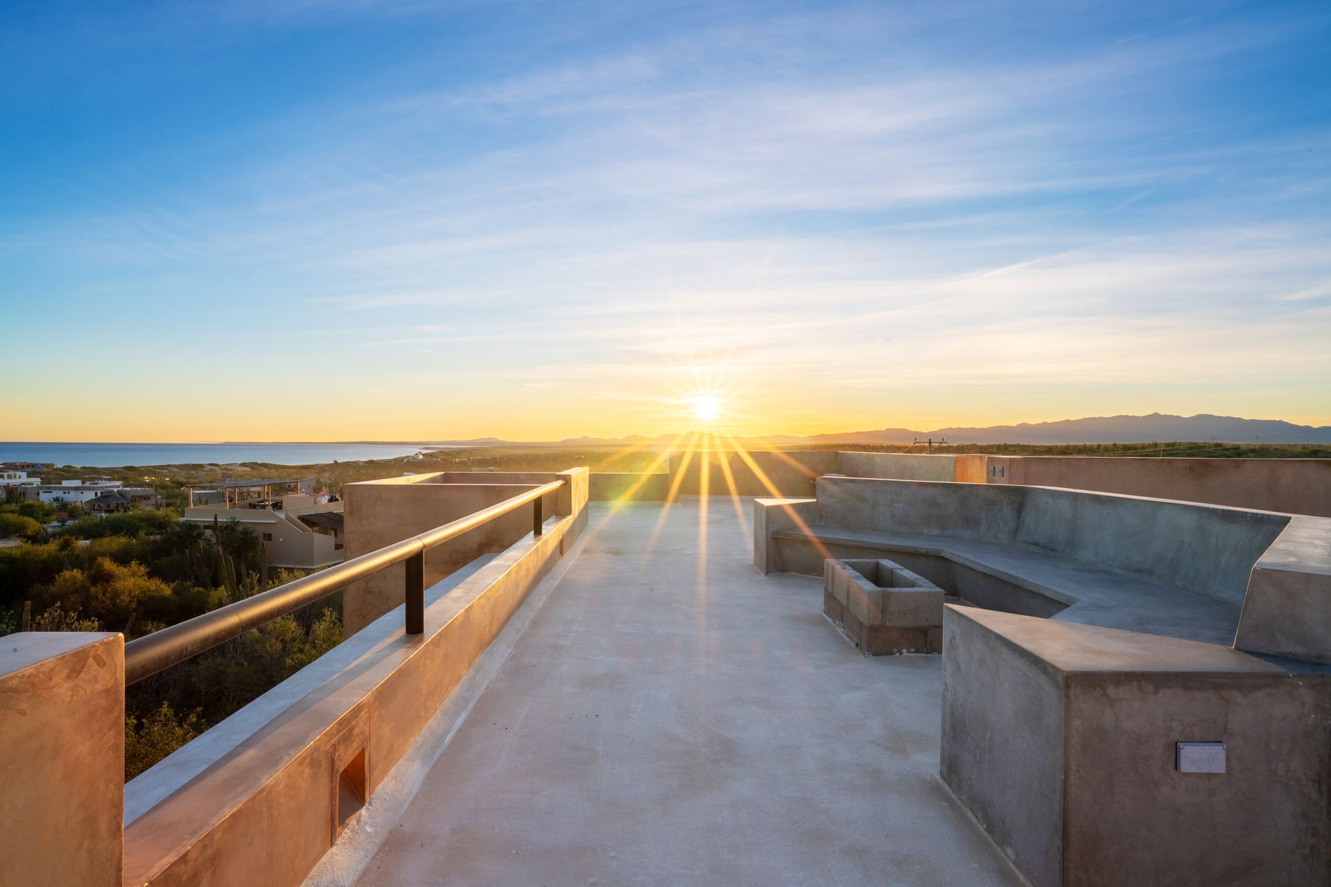 A sunset view from a concrete rooftop terrace featuring built-in seating, overlooking a landscape with trees and the sea.