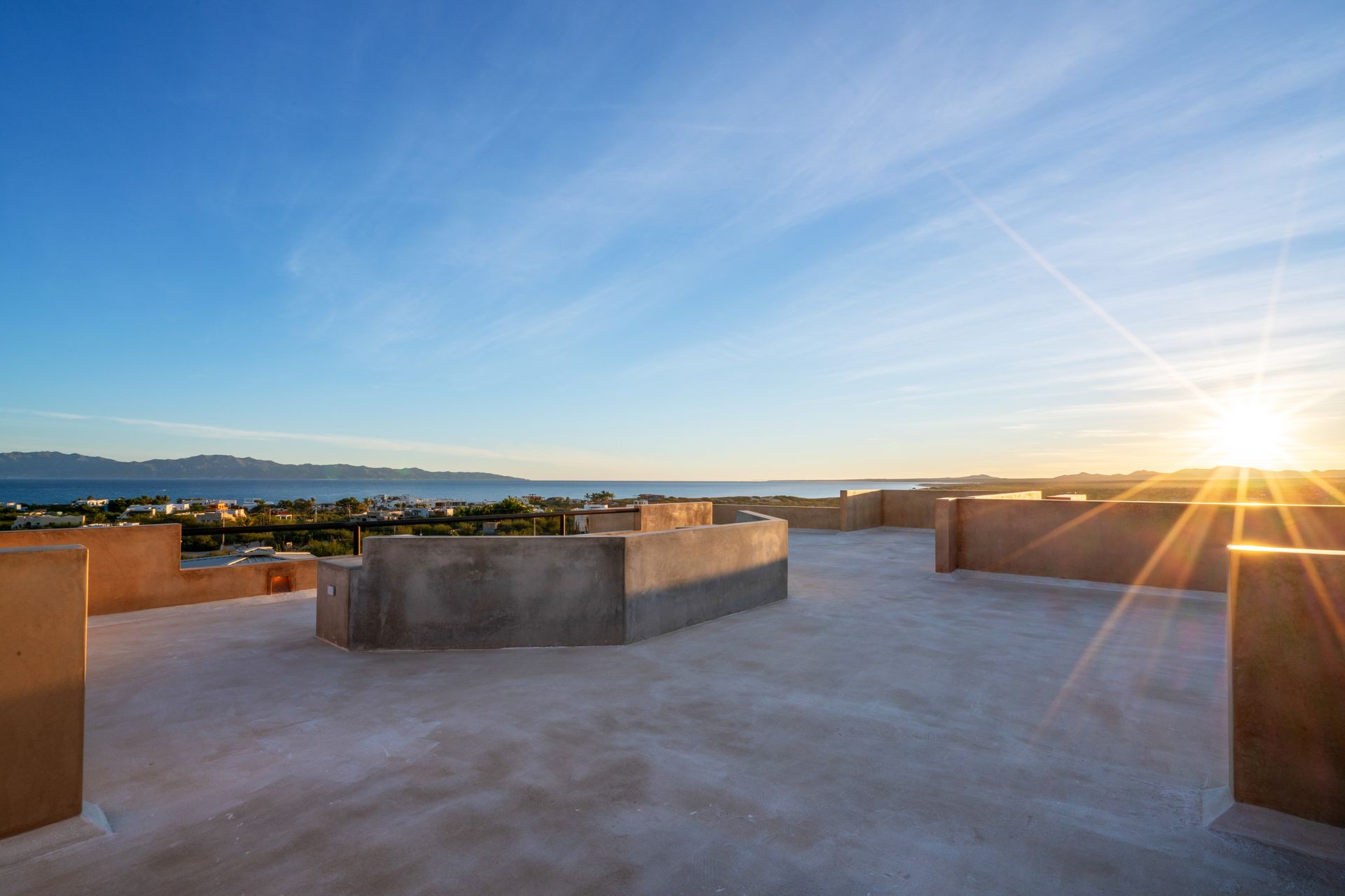 A rooftop terrace with stone walls overlooks a calm sea at sunrise under a bright blue sky.