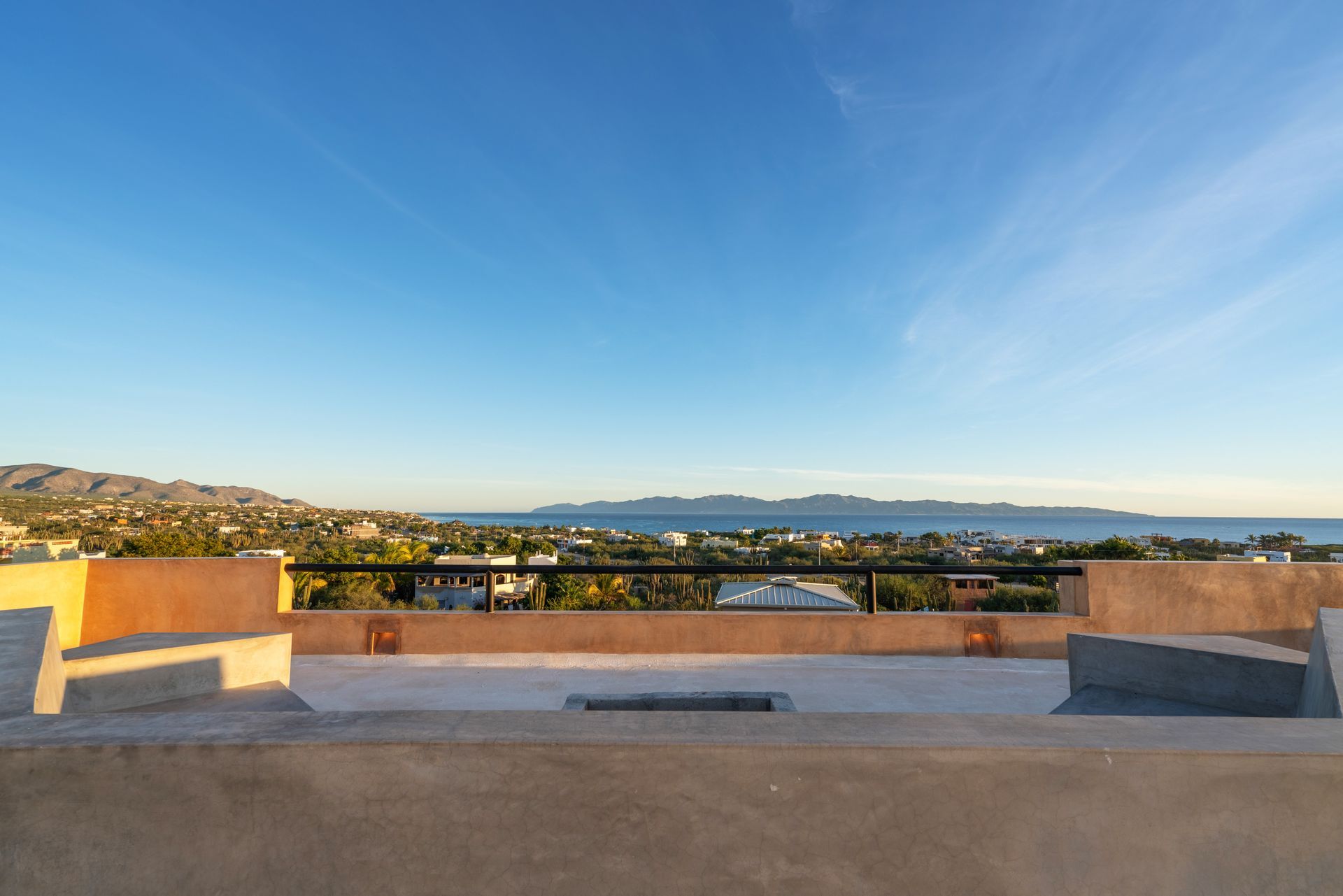 A panoramic view of the ocean and distant mountains from a terracotta-colored rooftop patio under a clear blue sky.