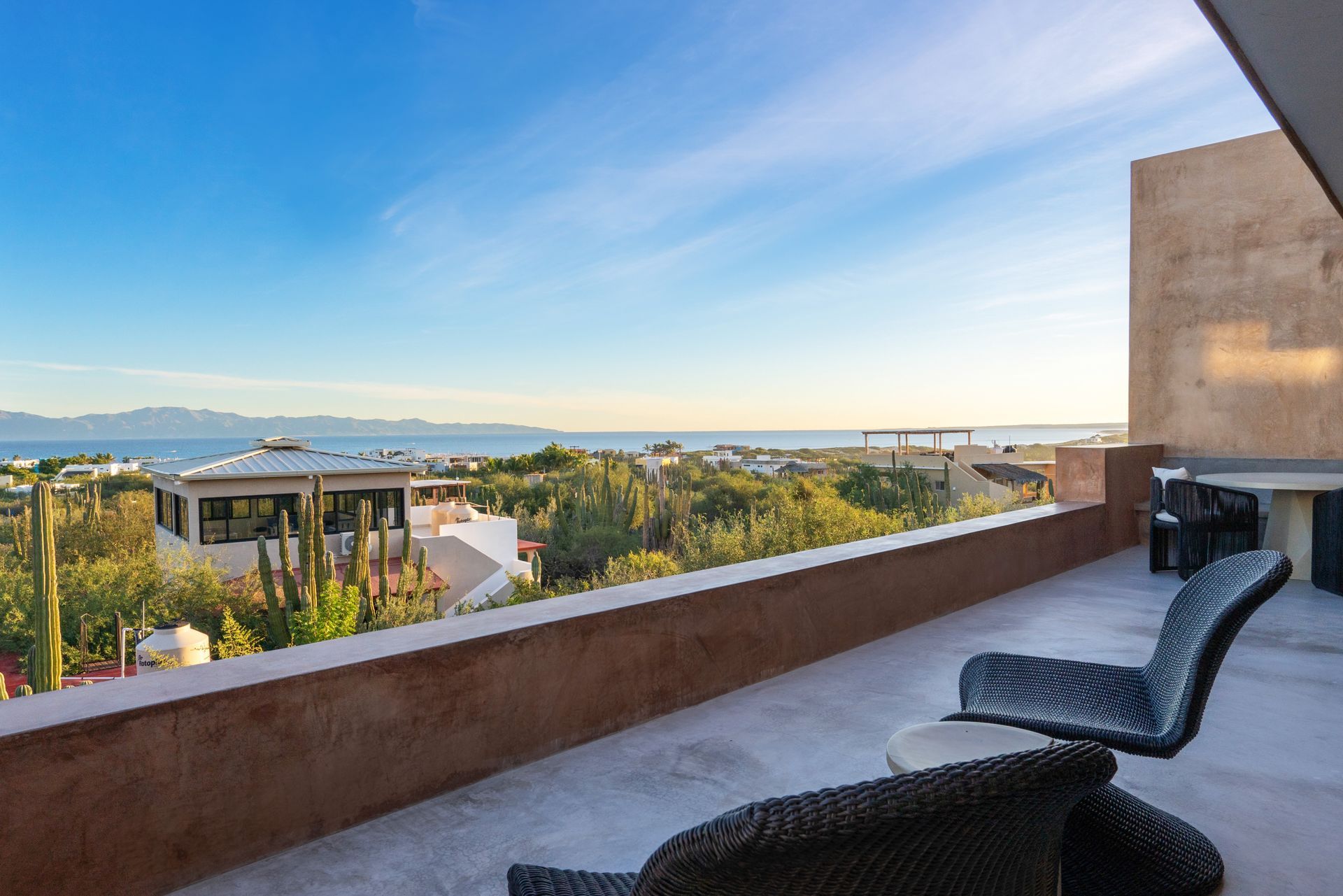 A sunny balcony overlooking lush vegetation and a distant ocean, with two dark wicker chairs in the foreground.