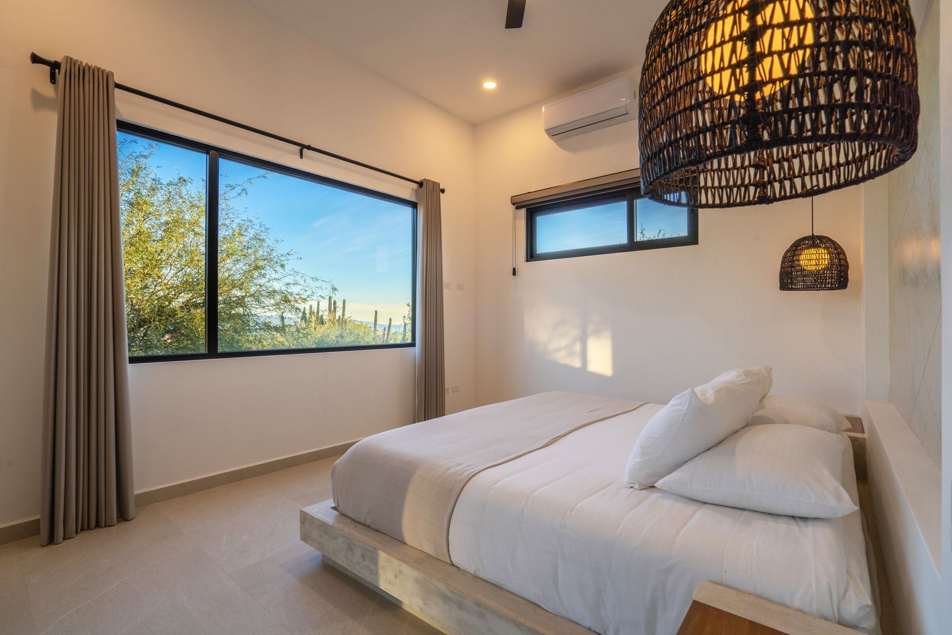 Minimalist bedroom with a platform bed, white bedding, large windows overlooking desert landscape, and a woven pendant lamp.