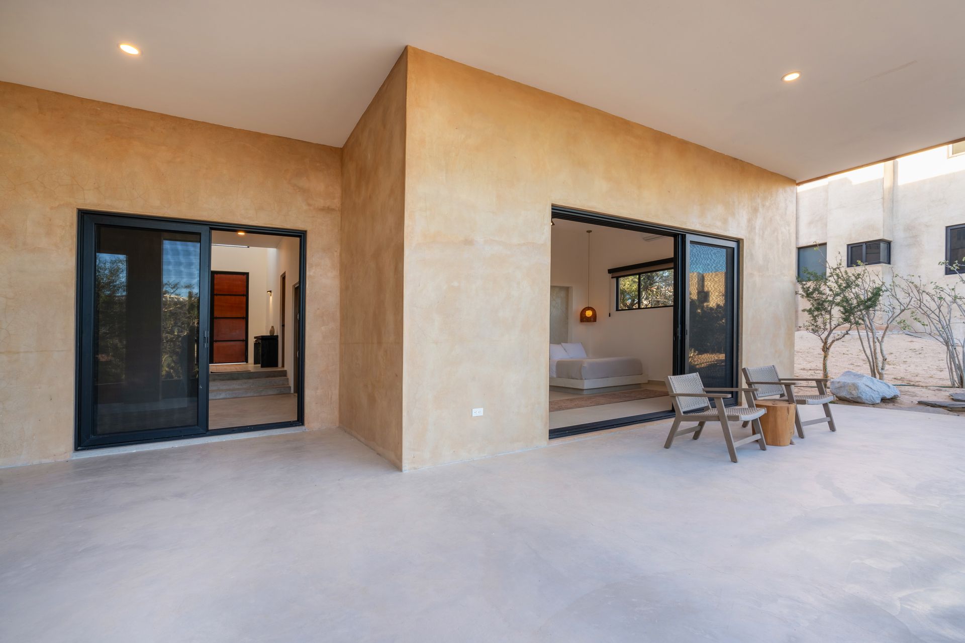 A beige, minimalist patio with two glass sliding doors opening into a house and two chairs placed on a concrete floor.