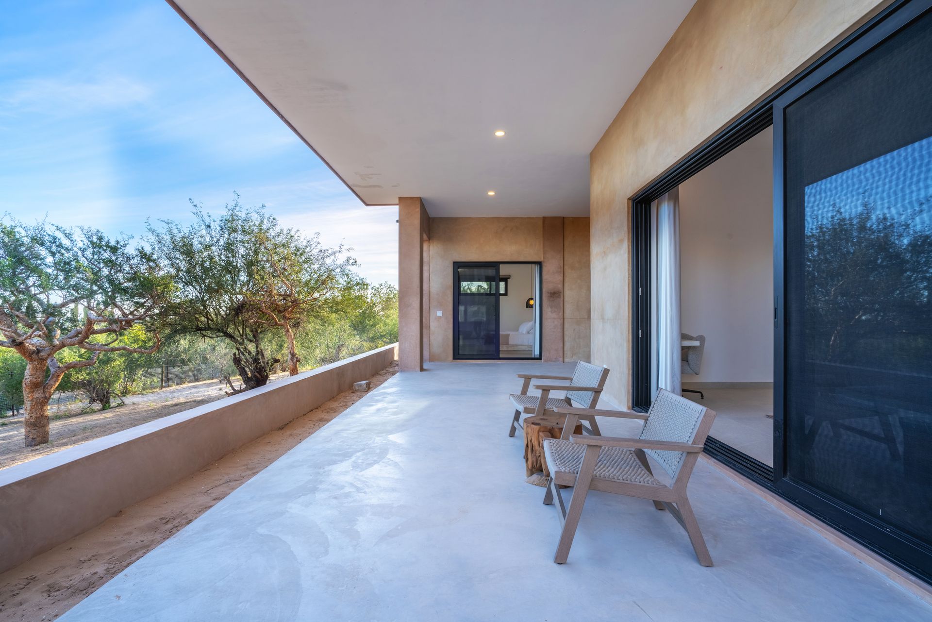 A wide, covered concrete patio features two wooden chairs, with sliding glass doors and outdoor trees in the background.
