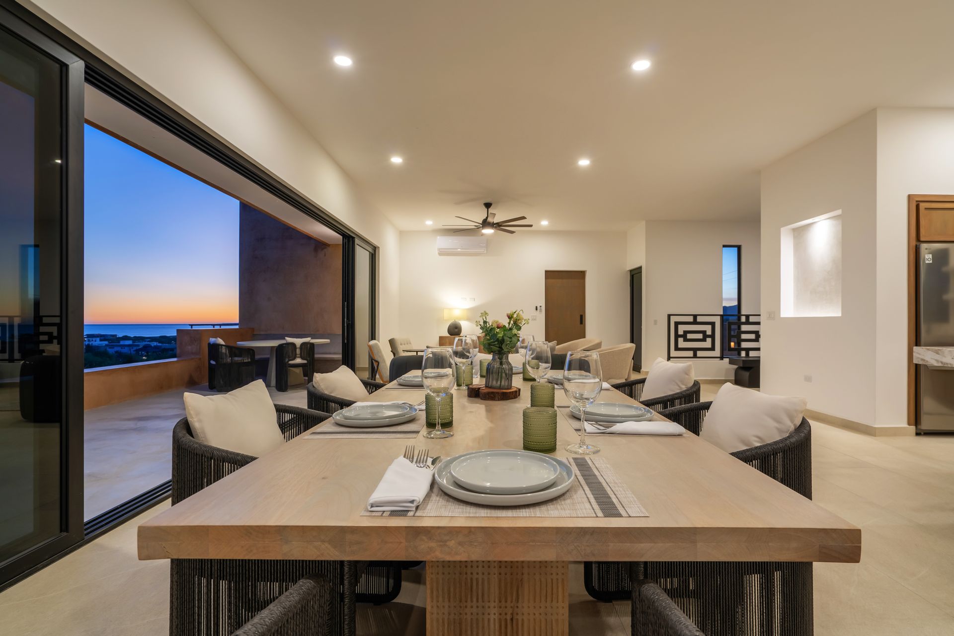 A dining table set for dinner in a modern, open-plan room with large sliding doors overlooking a sunset ocean view.