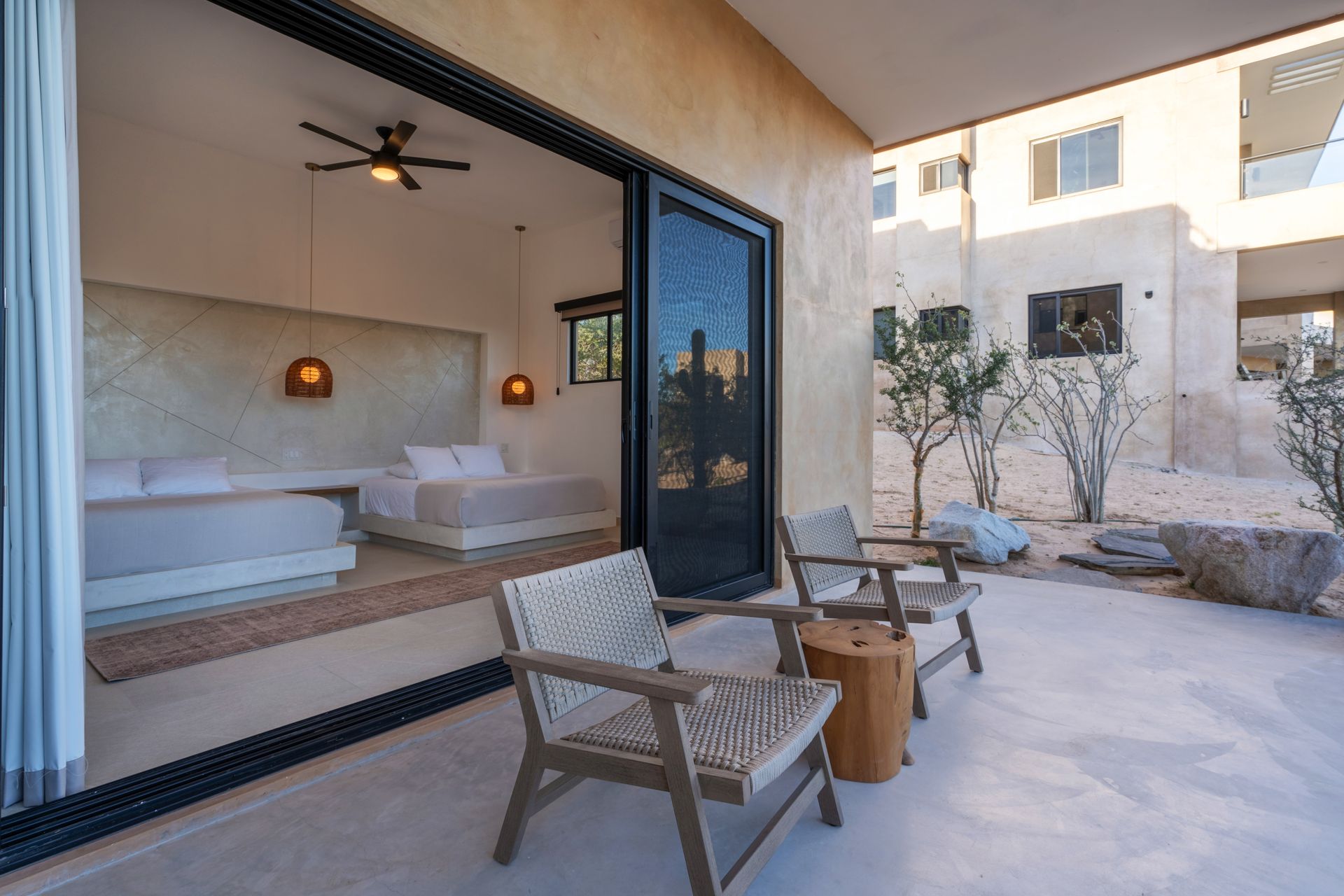 A minimalist bedroom with two beds opens onto a sunny patio with two chairs, wooden tables, and desert landscaping.