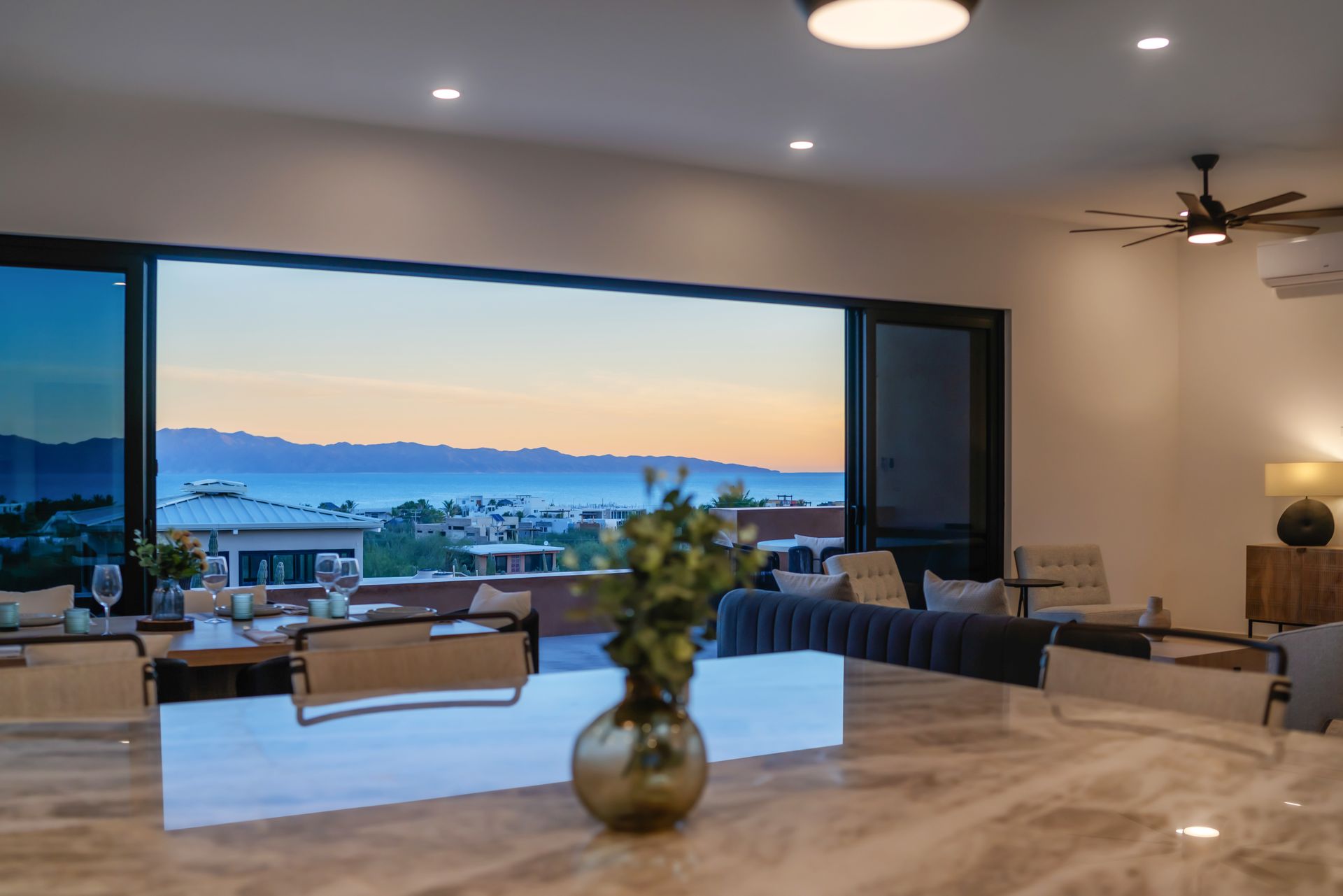A modern living area with an open-plan kitchen looking out onto a panoramic sunset view of mountains and the sea.
