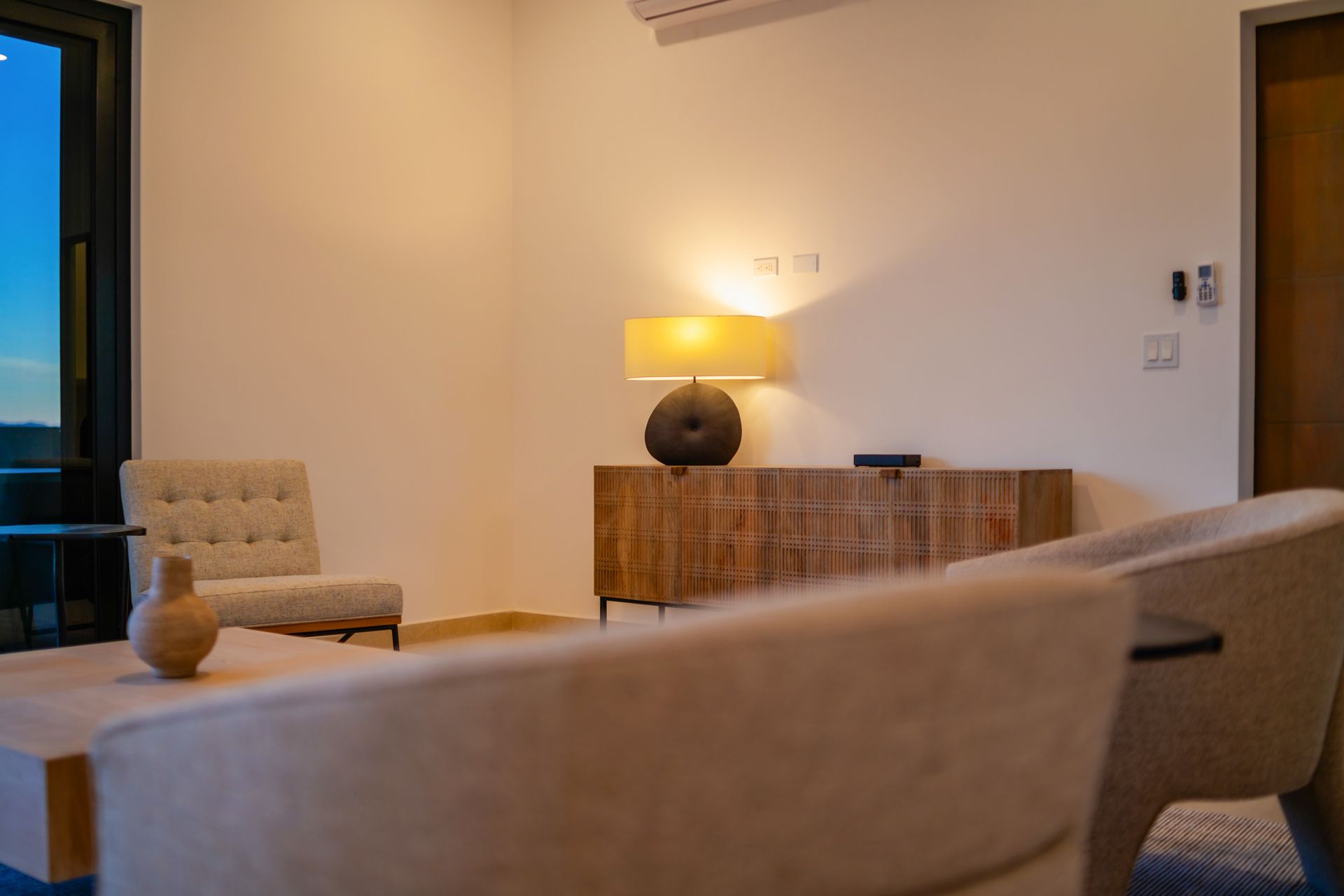 A modern, minimalist living room with beige armchairs, a wooden sideboard, and a warm glowing table lamp.