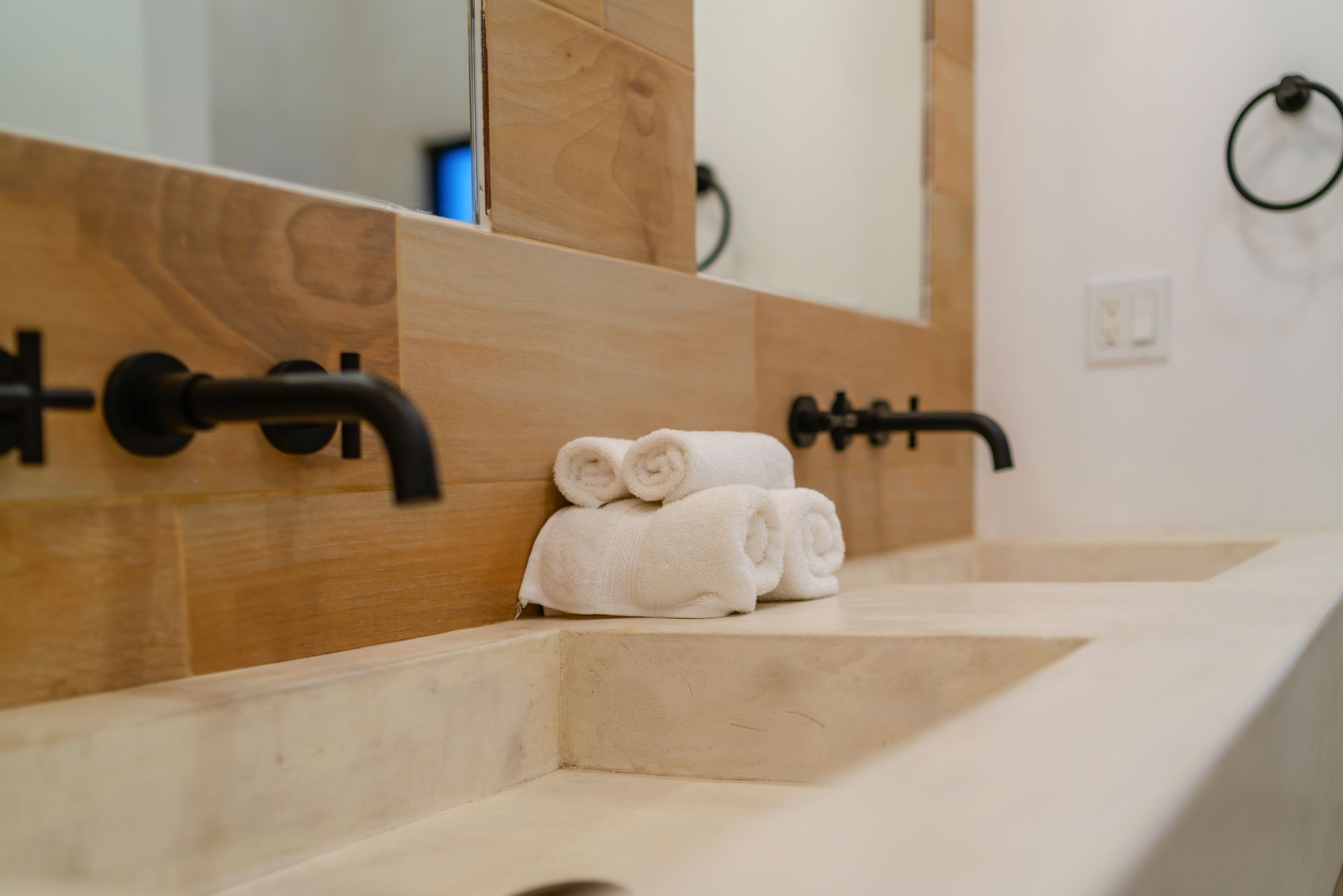 Double vanity with light wood backsplash, matte black wall-mounted faucets, stone sinks, and folded white towels.