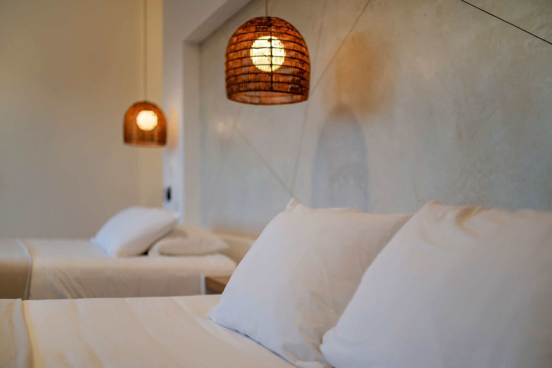 Two beds with white linens in a minimalist room, lit by woven hanging pendant lights against a textured light-gray wall.