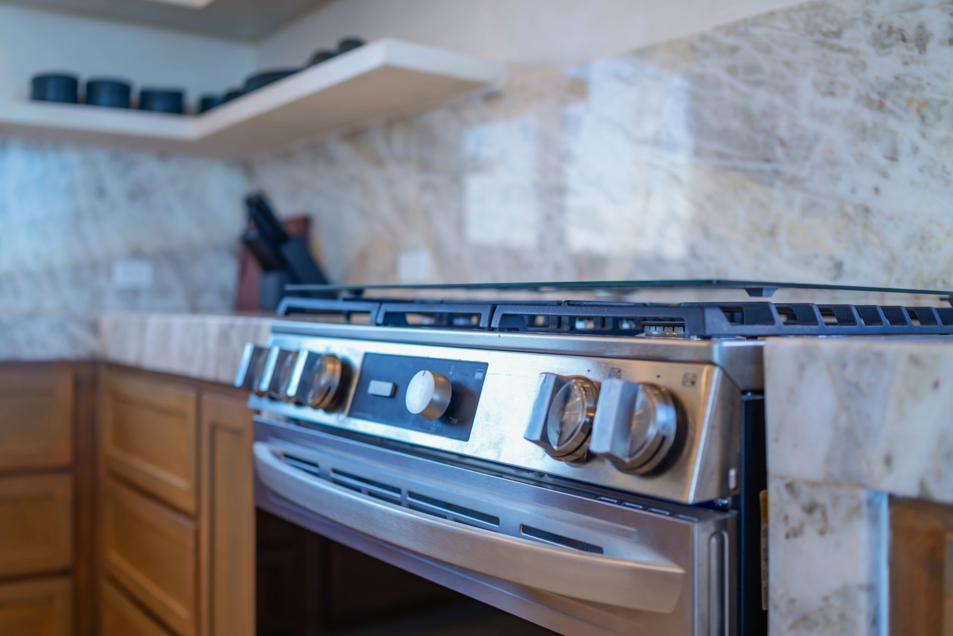 A stainless steel gas stove set into a stone countertop in a kitchen with wooden cabinets and a floating shelf.