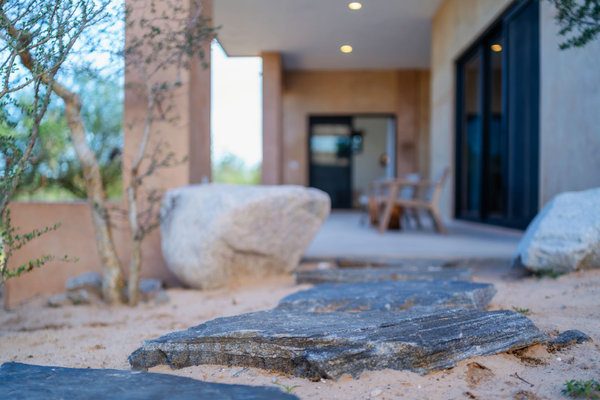 Flagstone stepping stones lead toward a modern patio with a large boulder and outdoor furniture in a desert landscape.