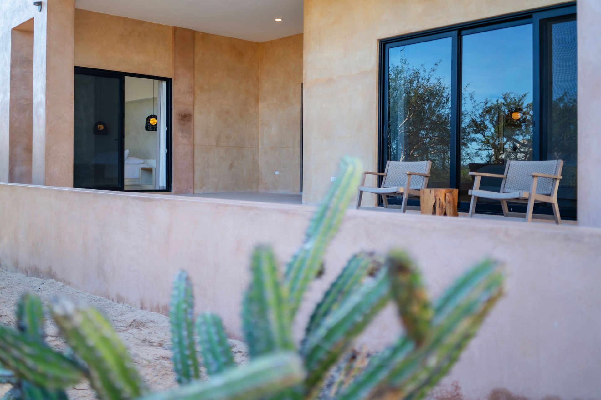 A patio with two chairs and a wooden stool, viewed past a green cactus, against a beige stucco building at dusk.