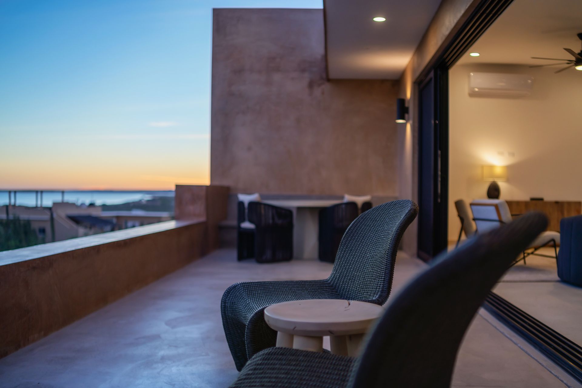 Wicker chairs on a modern terrace at sunset, with a view of the sea and a peek into an indoor living area.