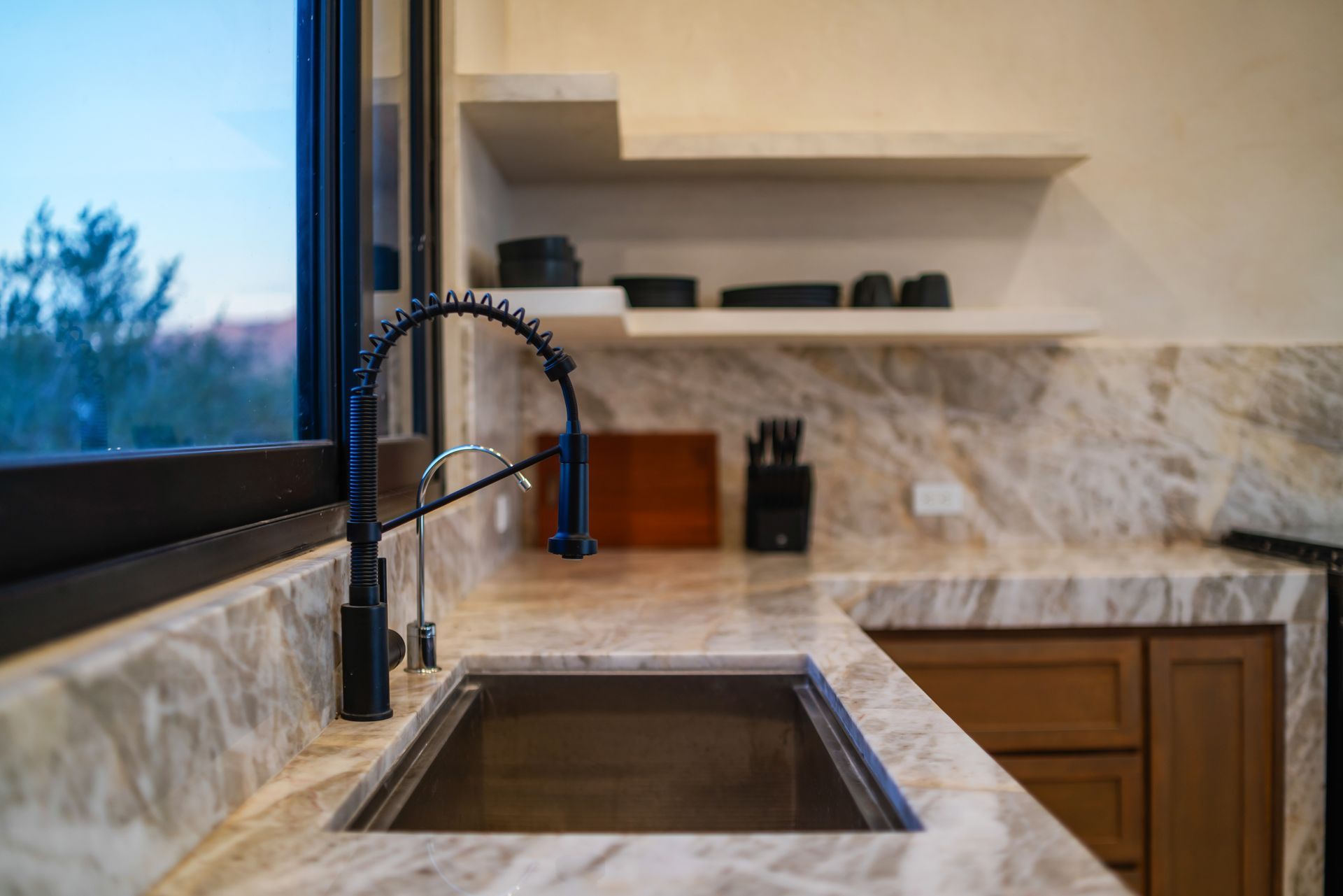 A modern kitchen sink with a black industrial faucet set in a light-veined marble countertop with wooden cabinets.
