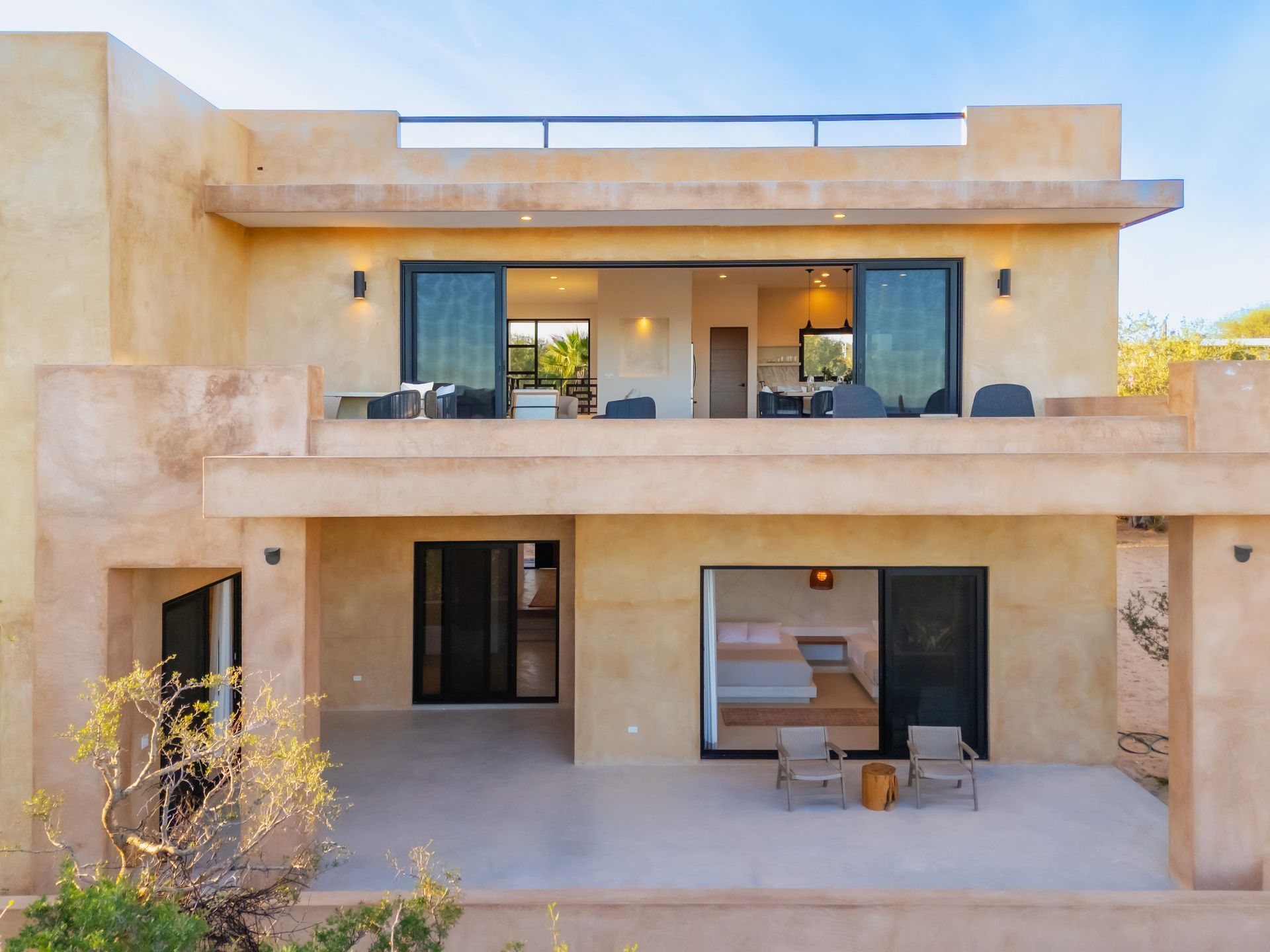 A two-story tan stucco villa featuring a ground-level patio and a large open upper balcony with dark sliding doors.