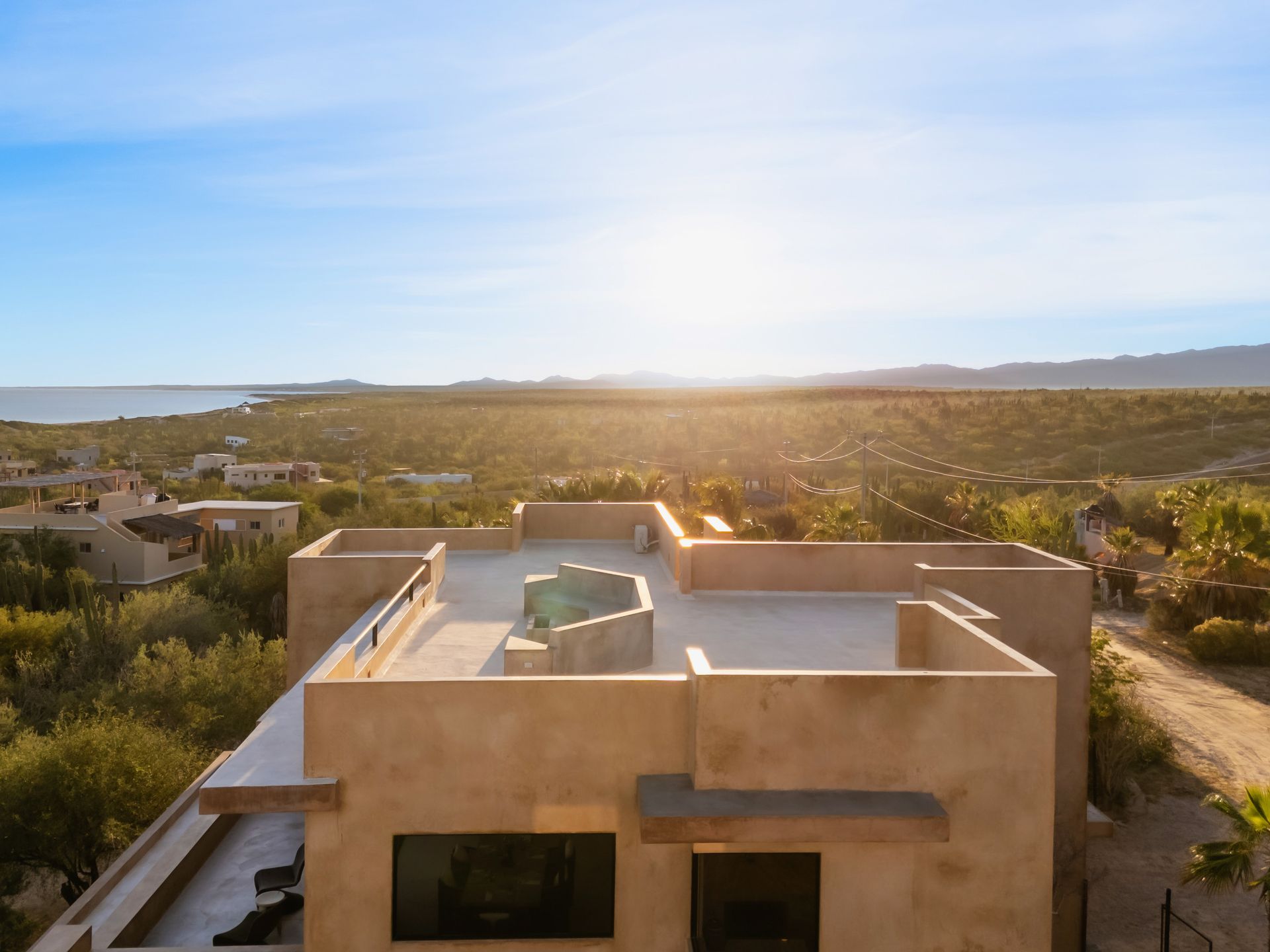 Aerial view of a tan, flat-roofed house in a desert landscape at sunset, with a sea horizon in the distance.