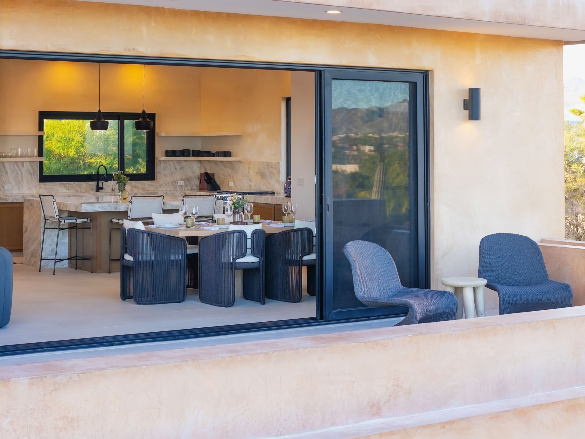 Modern patio dining space transitioning to an open-plan kitchen, with blue wicker chairs, stone walls, and scenic views.