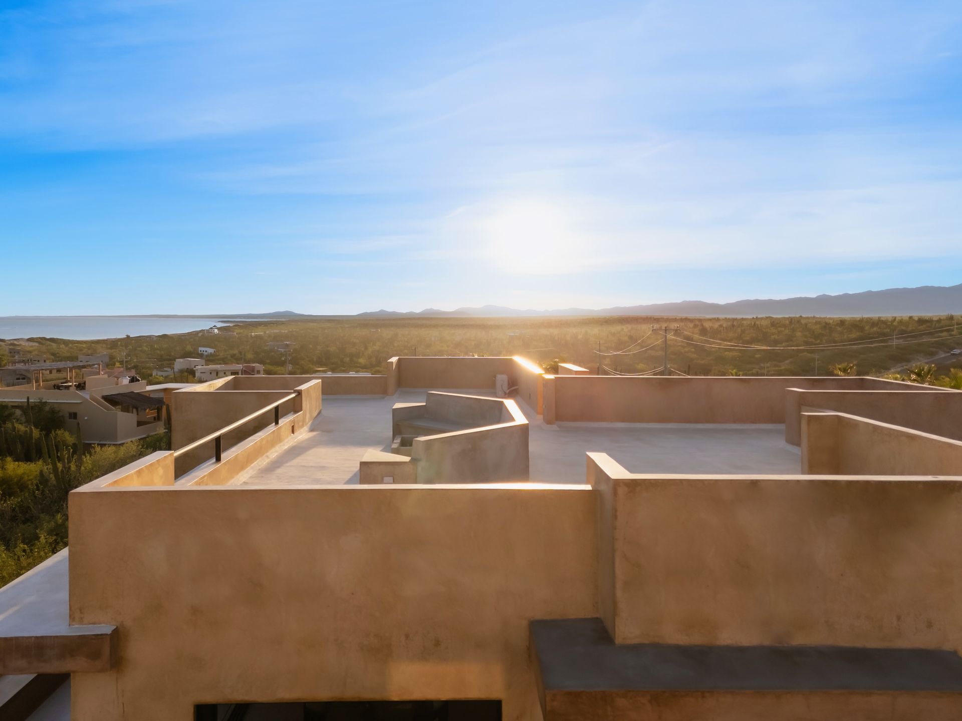 A sun-drenched rooftop terrace with tan stucco walls, overlooking a distant desert landscape and the ocean.