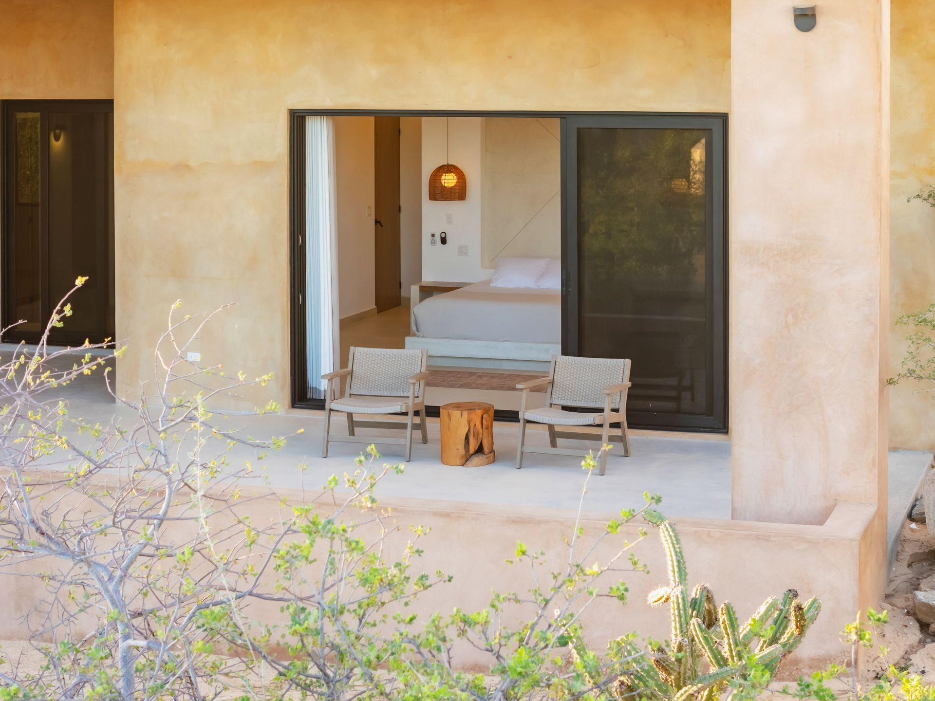 Two chairs and a wooden side table on a patio, overlooking an open bedroom door with a bed inside in a desert setting.