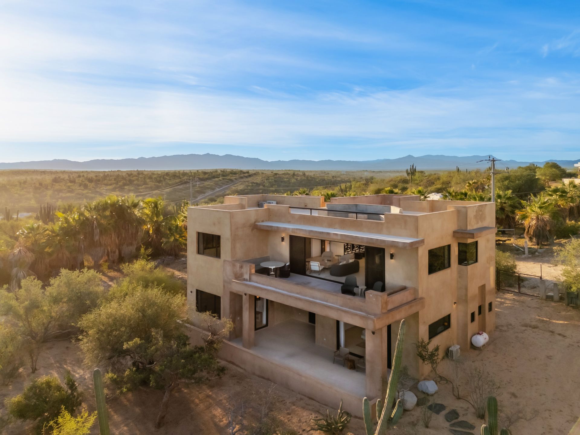 A two-story, flat-roofed desert villa with beige stucco walls, surrounded by trees and cacti under a sunny blue sky.
