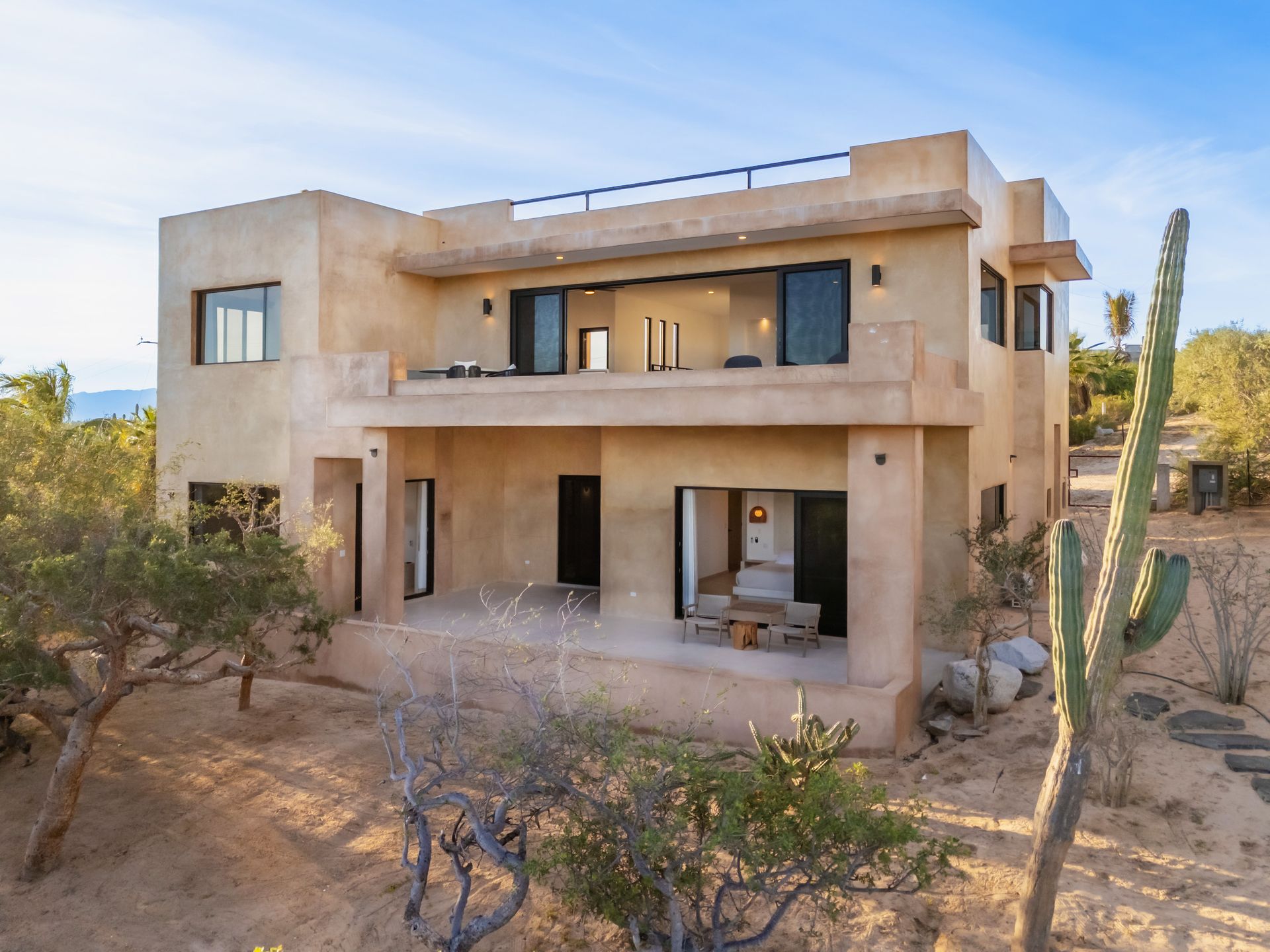 A two-story, tan-colored desert villa with a balcony, open-air patio, and surrounding cactus landscape under a blue sky.
