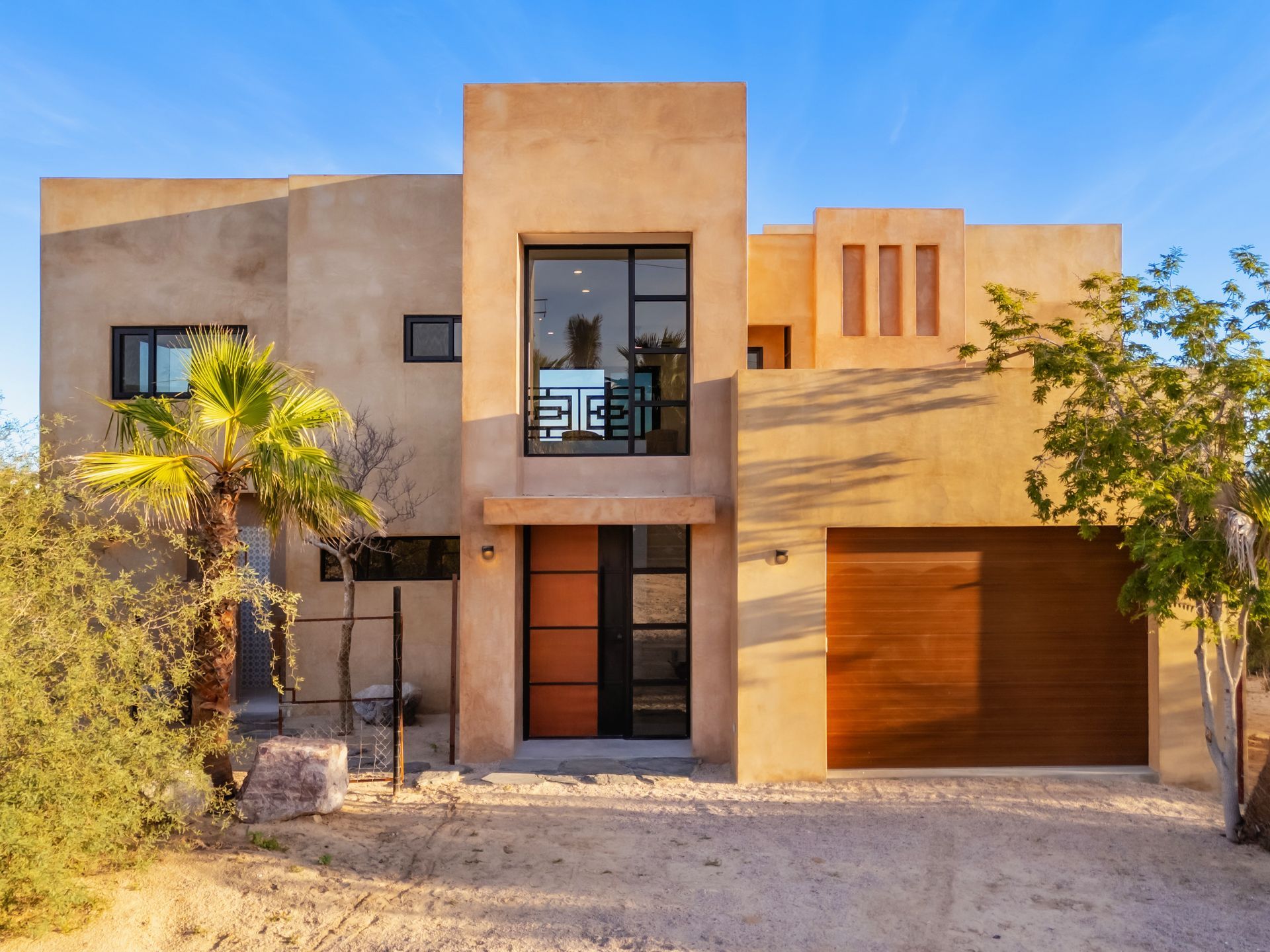 A two-story tan stucco house with a modern wood-paneled garage door and a front entrance, set in a desert landscape.