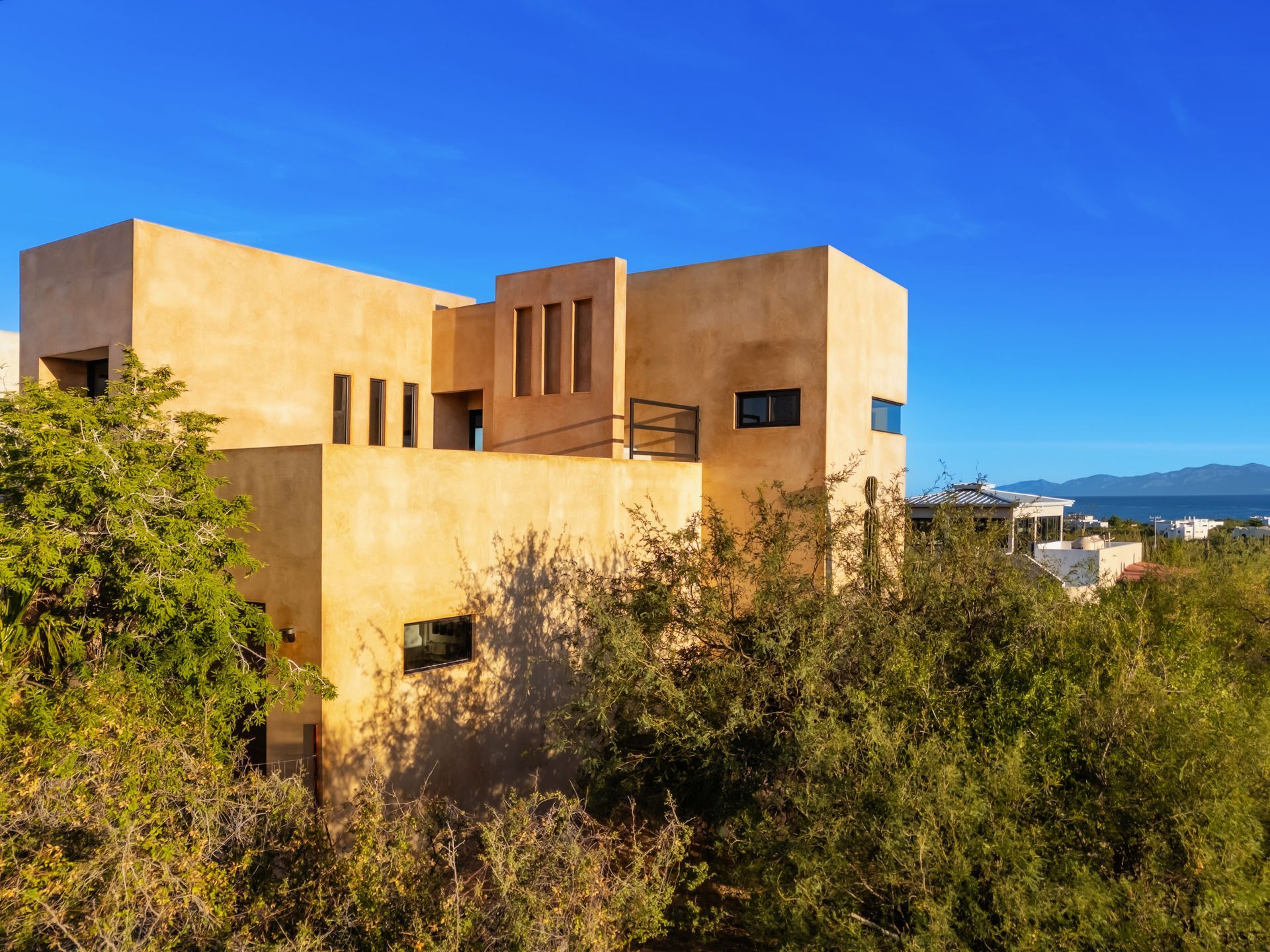 A tan, flat-roofed modern house sits amid lush green trees under a bright blue sky, with a distant view of the sea.