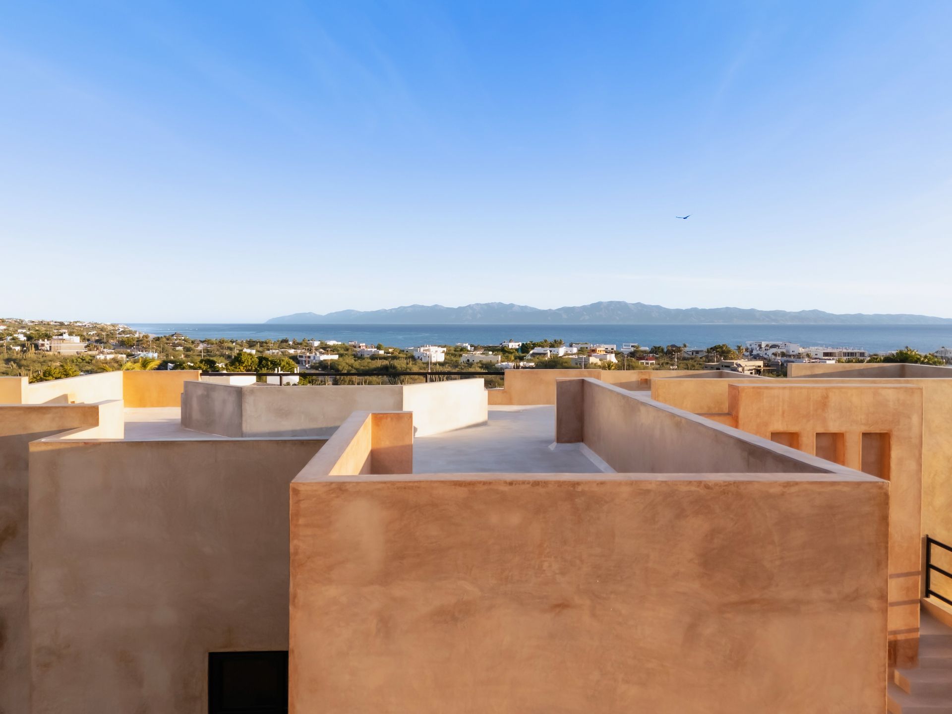 A flat-roofed, tan-colored structure overlooks a coastal town and sea with a distant mountain range under a clear sky.